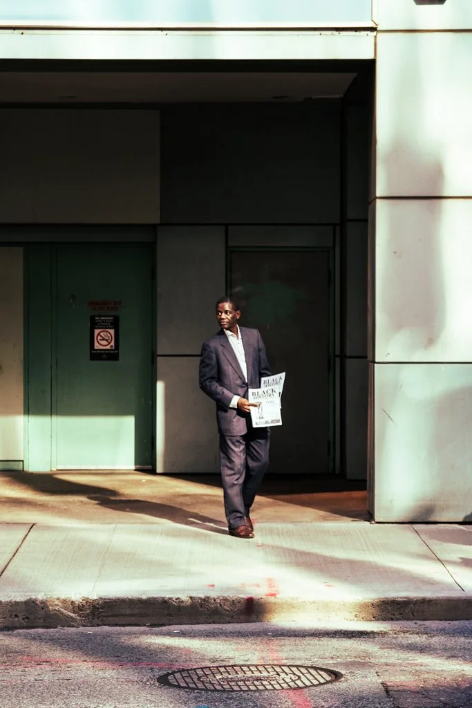 A man in a suit walking on the sidewalk, holding a newspaper, near a building with concrete walls and a green door.