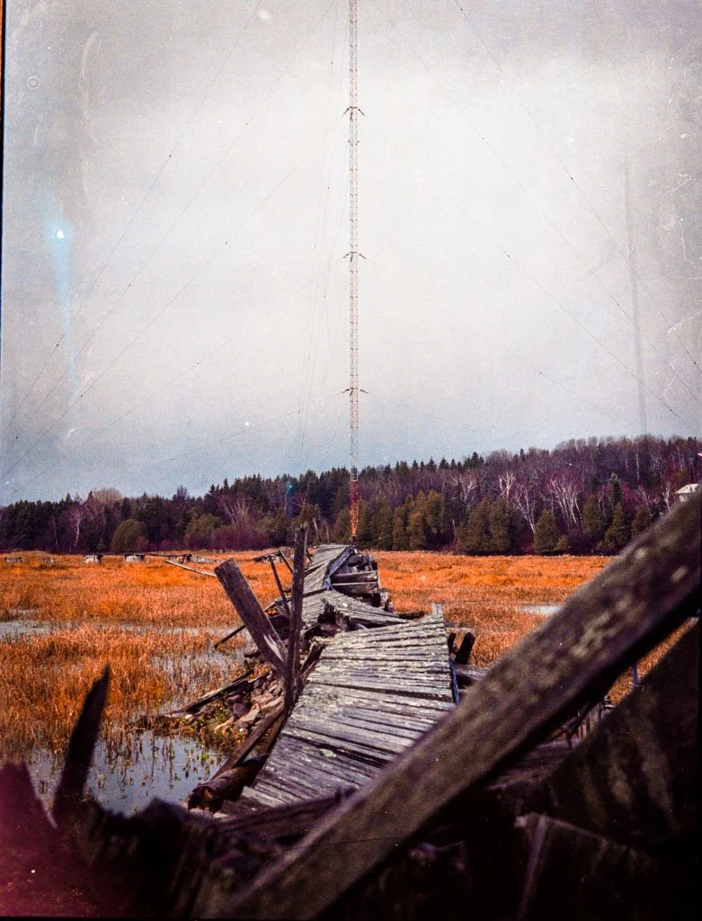 A broken, dilapidated wooden bridge leading to a field with marshy water, surrounded by trees and under an overcast sky, with a tall radio or communication tower in the background.