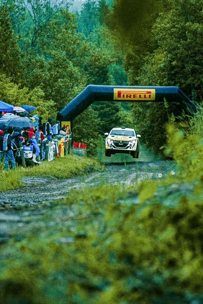 A rally car racing on a muddy forest rally stage with spectators watching from the side and a Pirelli banner overhead.