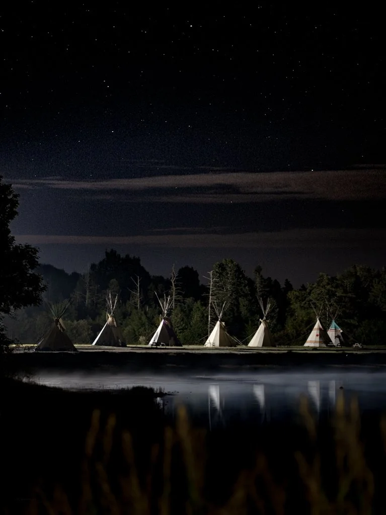 Nighttime scene of several teepees lined along the edge of a lake, with a star-filled sky overhead and trees in the background.
