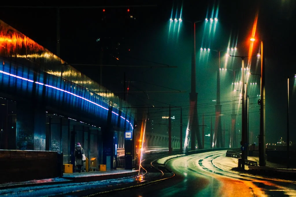 Nighttime scene of a deserted city street with wet road, illuminated streetlights, and a bridge structure in the background, with a person standing at a bus stop.