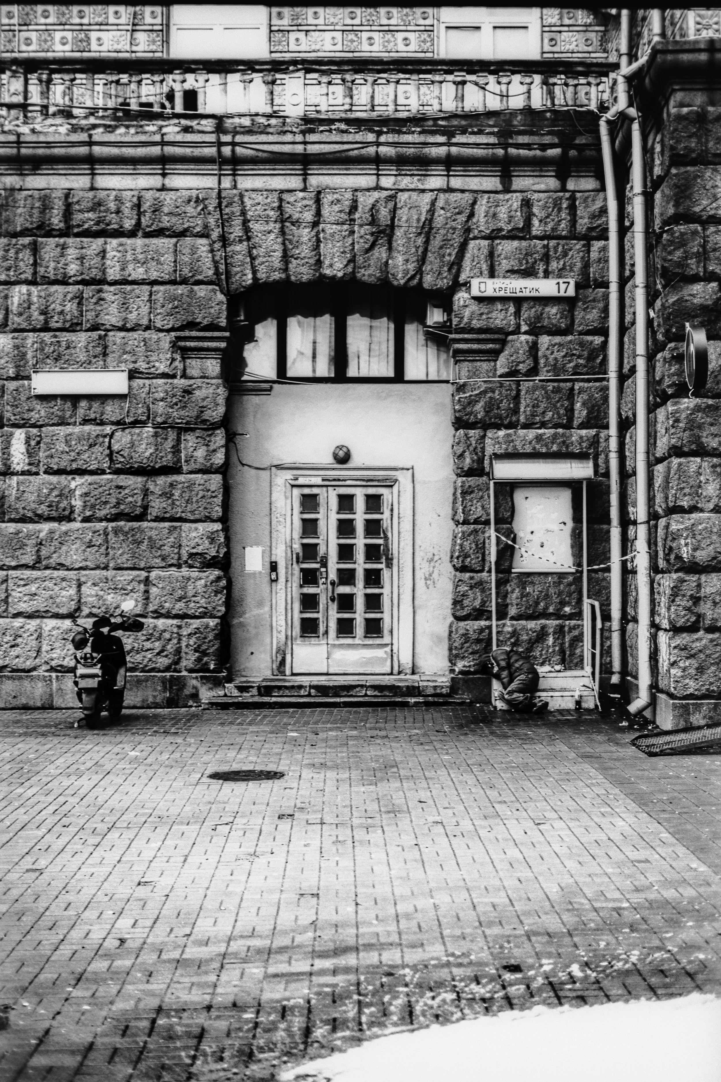Black and white photo of an old stone building with a large window above a door, street sign with Cyrillic text, scooter parked to the left, and a person crouched near the right side of the door.