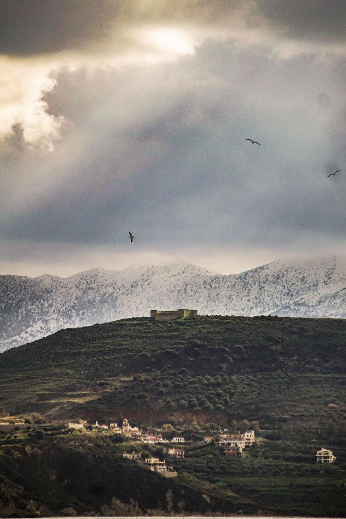 Mountain landscape with a structure on top, houses in the foreground, cloudy sky, and three birds flying.