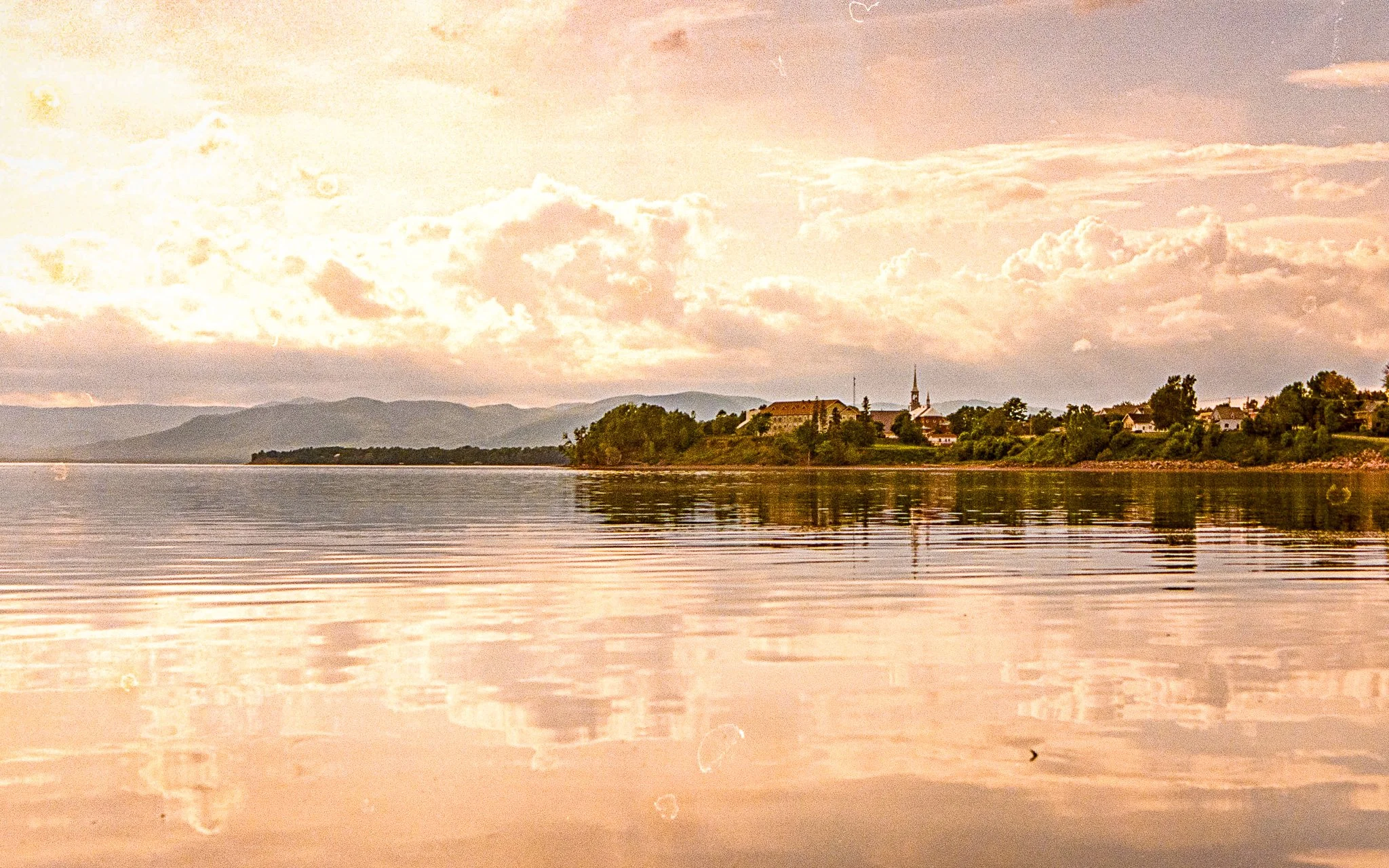 A calm lake with gentle ripples, reflecting the sky and clouds above. A small town with a church steeple is visible on the shoreline, with mountains in the background during sunset or sunrise.