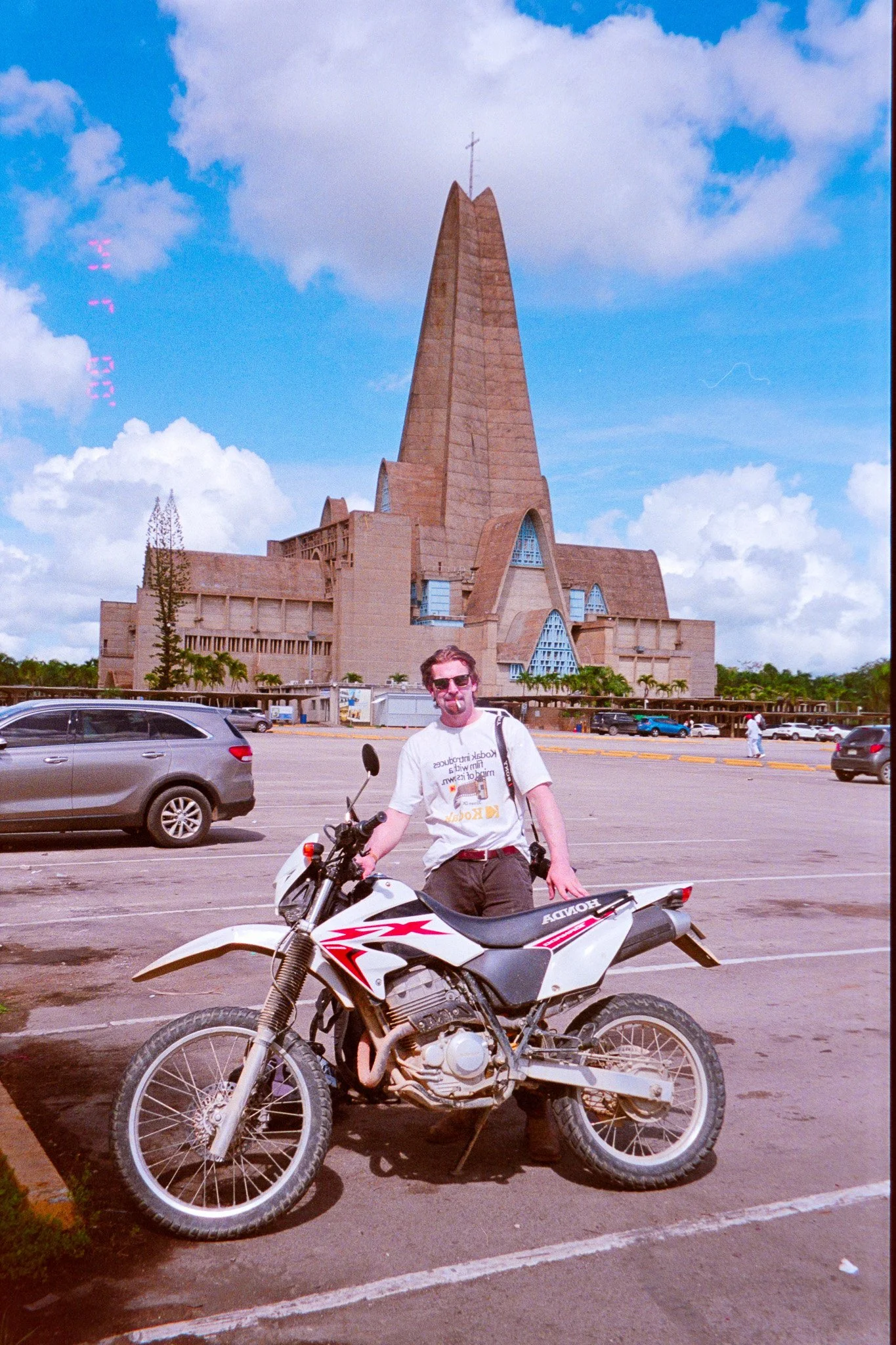 A man with sunglasses and a white T-shirt standing beside a white off-road motorcycle in a parking lot, with a large modern church building featuring a tall, pointed spire in the background.