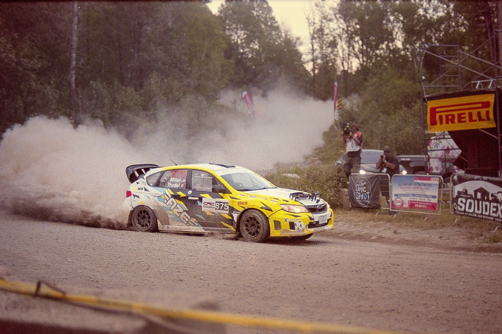 A yellow rally car speeding on a dirt road during a race, kicking up dust behind it, with spectators and banners on the side and a forest in the background.