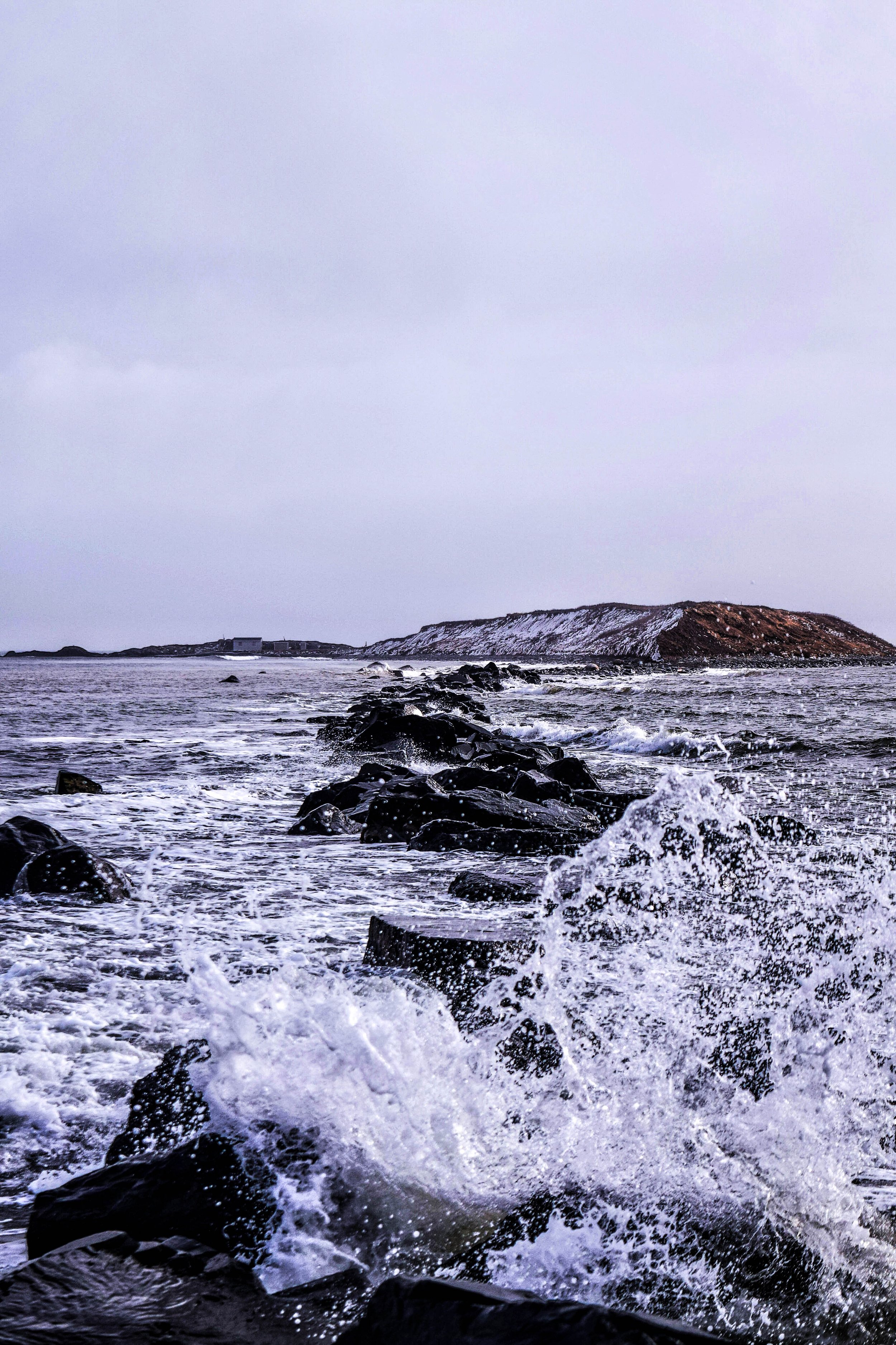 Ocean waves crashing against dark rocks with a snow-covered mountain in the background under an overcast sky.