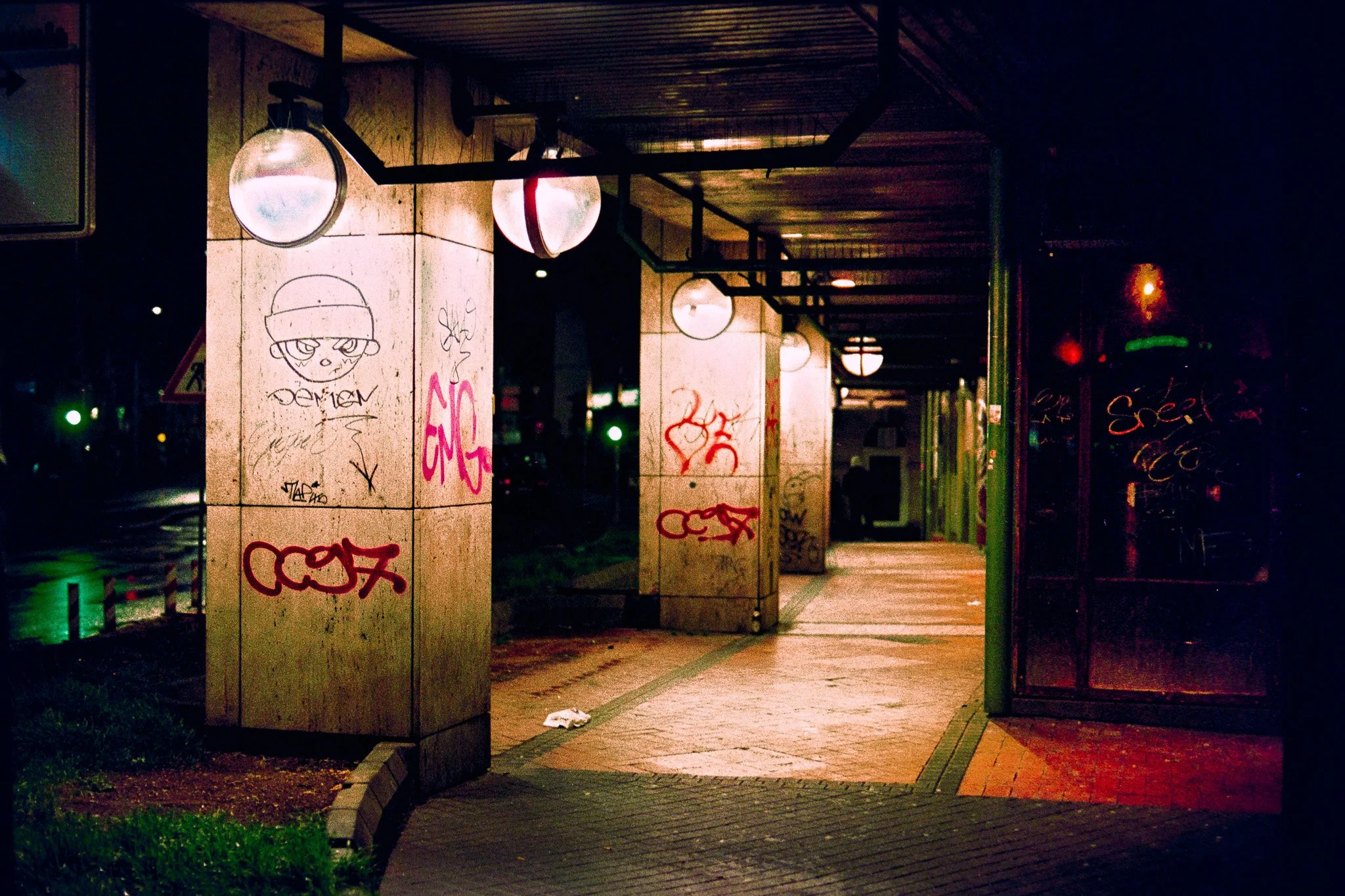 Empty sidewalk at night with graffiti-covered pillars and a glass bus stop shelter reflecting the street lights.