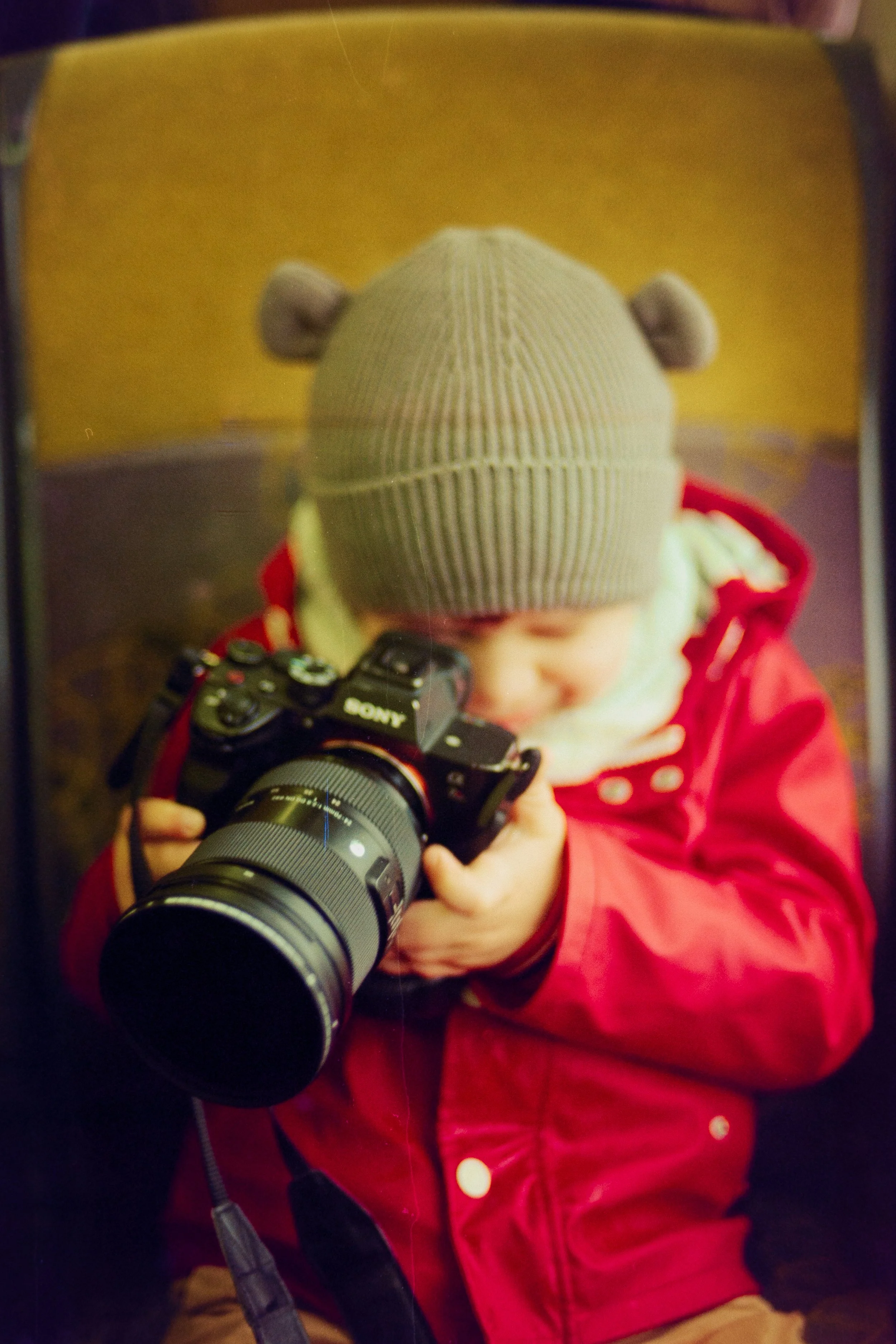 A young child in a gray knit hat with bear ears and a red jacket is looking at a Sony camera, smiling.