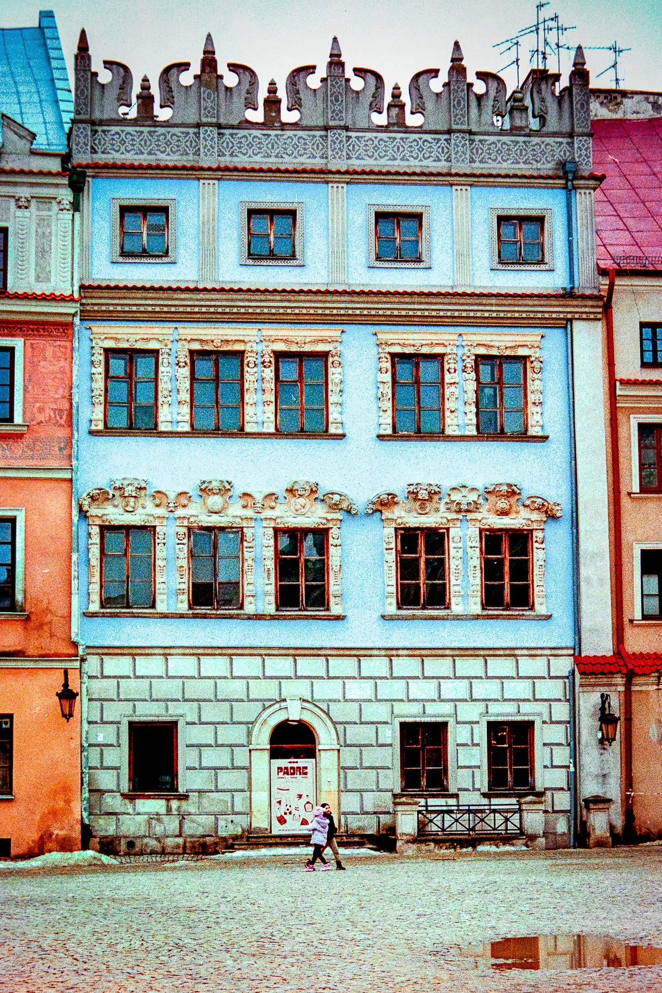 Colorful multi-story building with ornate window frames and decorative roofline, two children walking in front, cobblestone ground, and adjacent buildings.