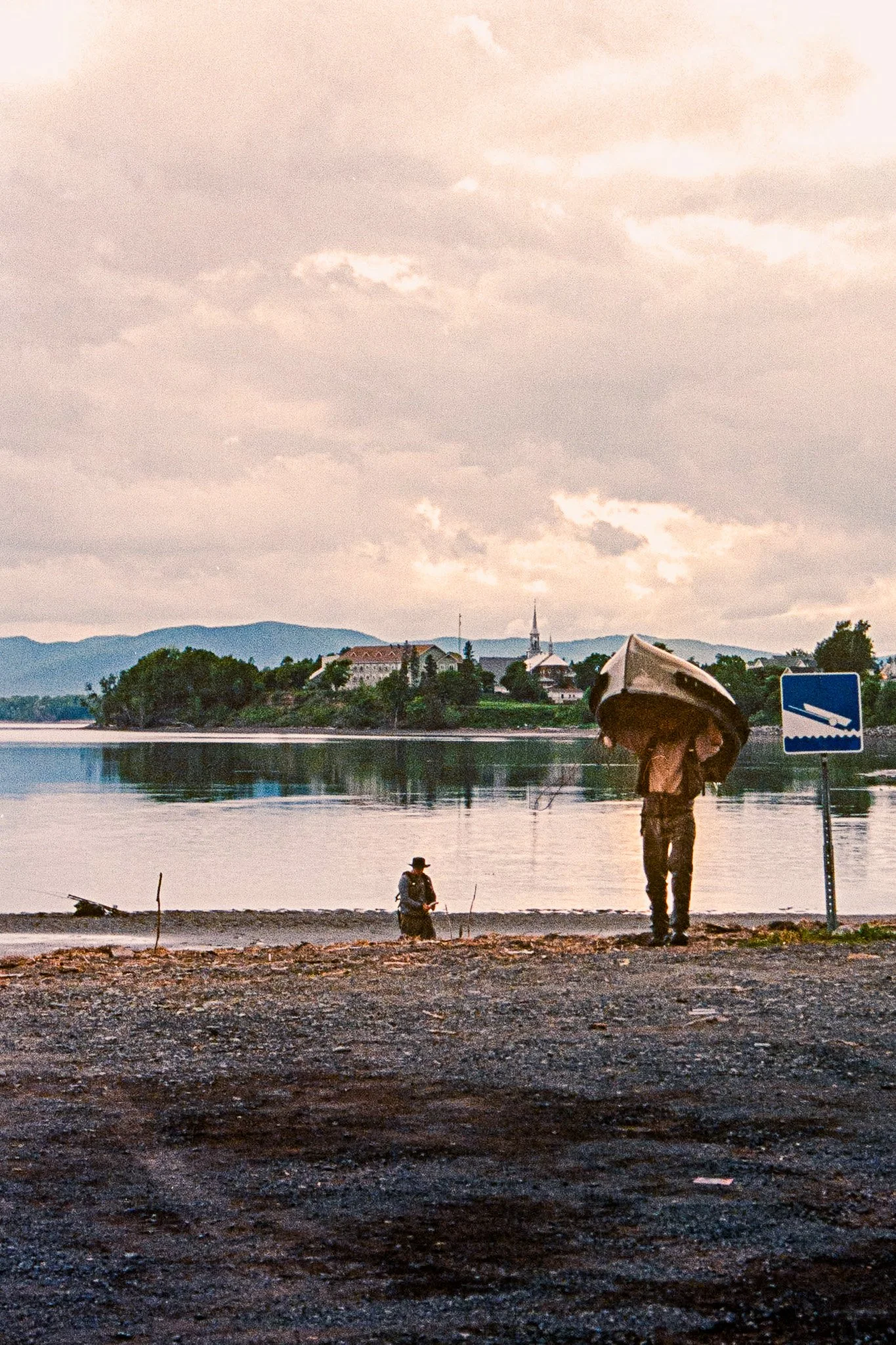 Two people by a riverbank, one standing holding an umbrella and the other sitting with a hat, with a town and mountains in the background.