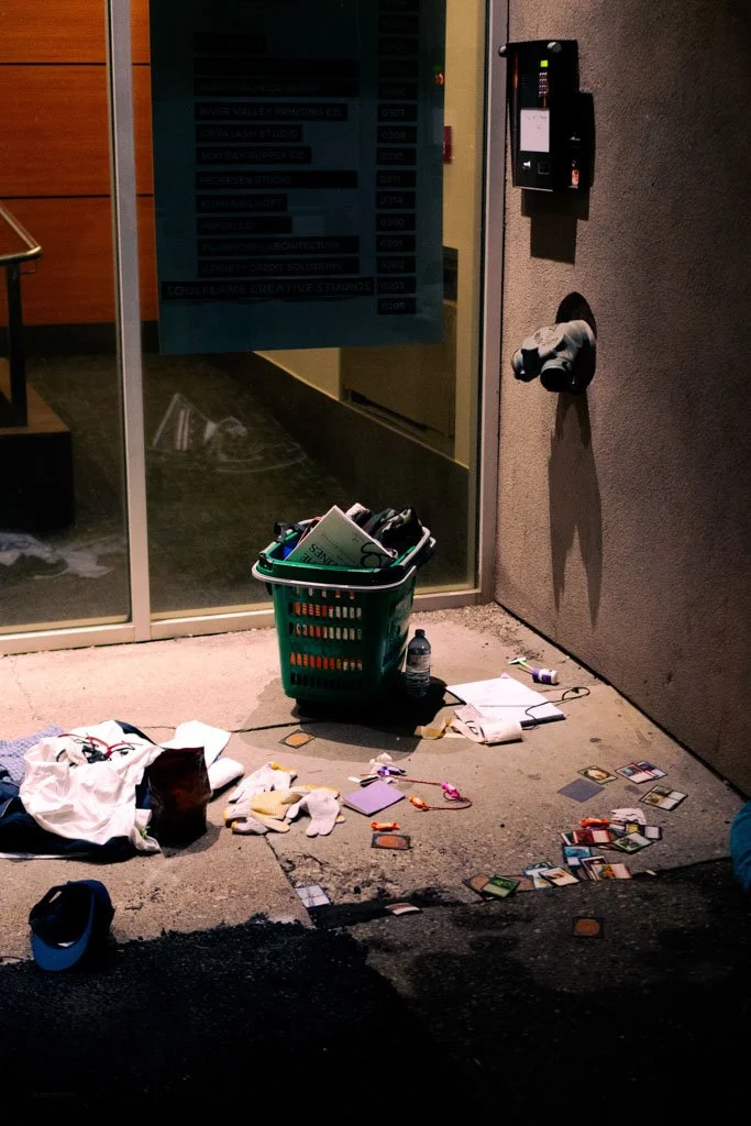 A messy outdoor scene with a laundry basket filled with papers, a water bottle, and scattered small items on the ground. Nearby are gloves, a piece of cloth, and playing cards. There's a glass door with a sign inside, a mailbox mounted on the wall, a