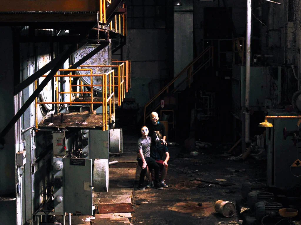 Three people with masks in a dark, industrial setting with pipes, stairs, and machinery.