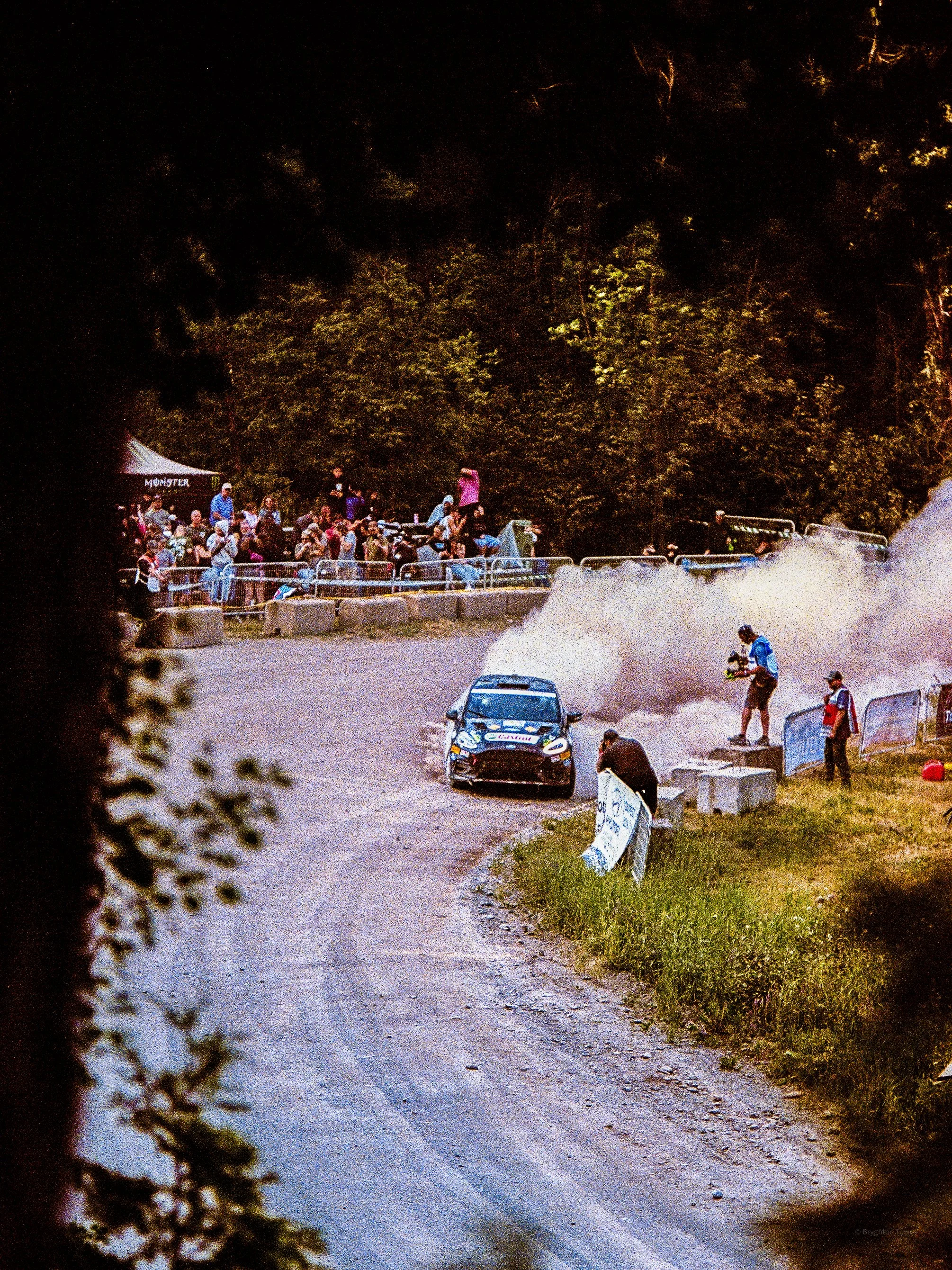 A rally car race taking place on a dirt road, with a car kicking up dust and spectators watching from the side