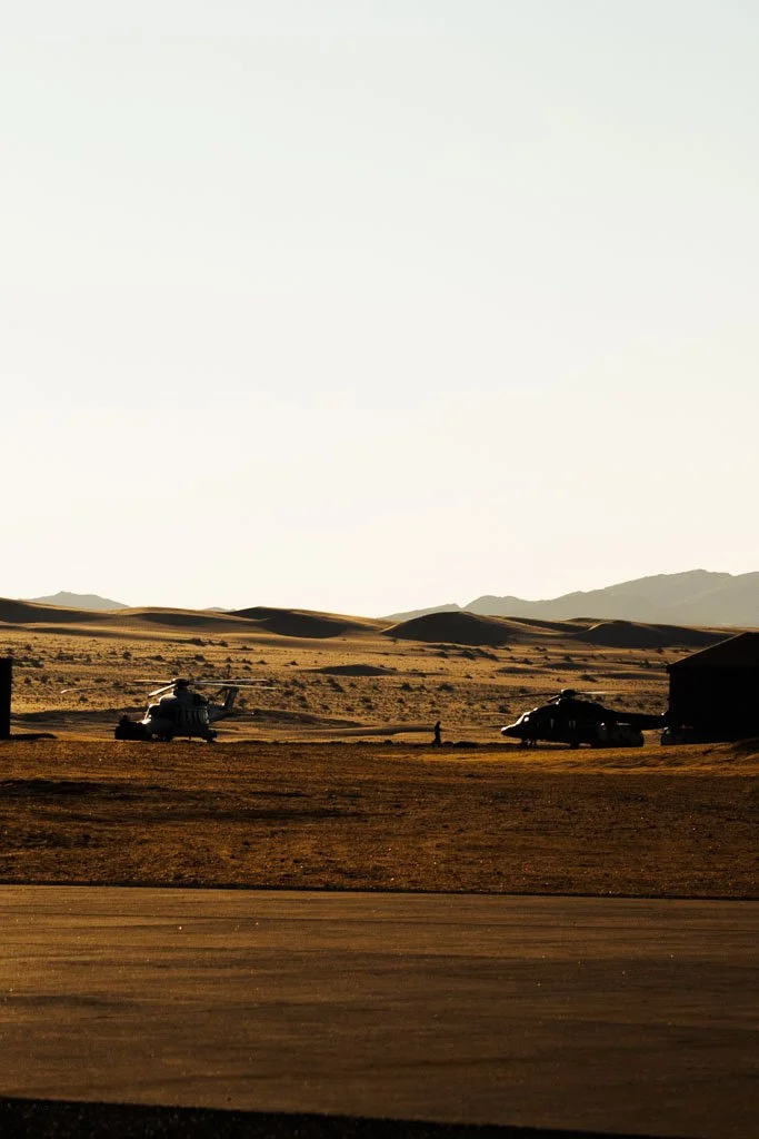 Desert landscape at sunset with two helicopters and a vehicle on the ground.