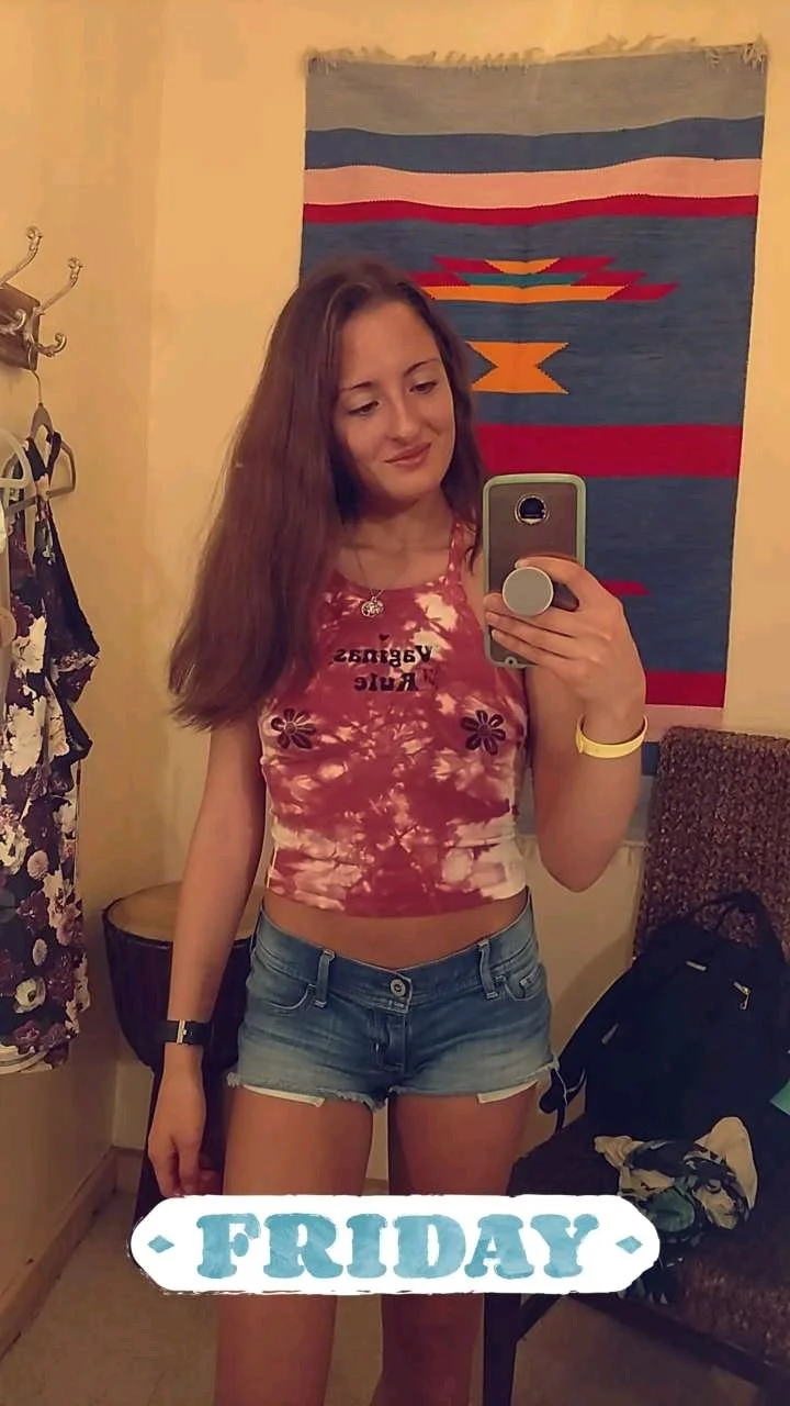 Jenny taking a mirror selfie wearing a tie-dye tank top and denim shorts, with a tapestry hanging in the background.