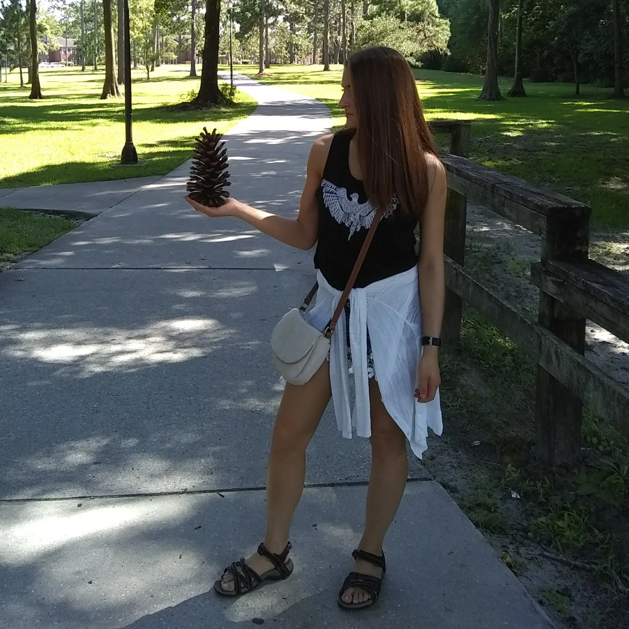 Jenny standing on a path in a park, holding a large pine cone in her hand.
