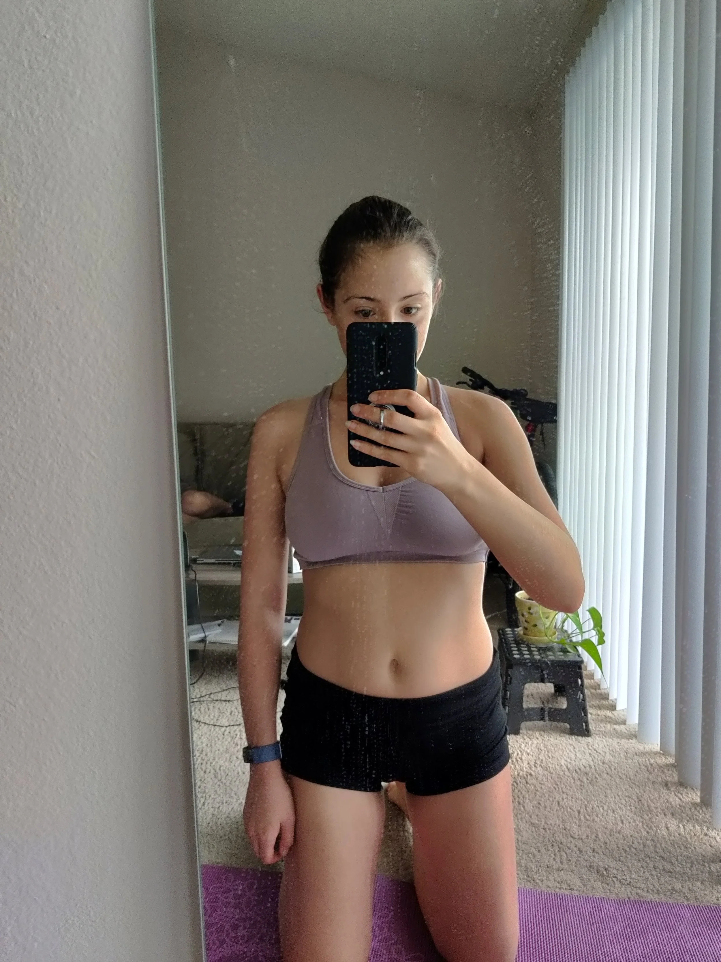 Jenny taking a mirror selfie wearing workout clothes inside a room with carpeted floor and vertical blinds.