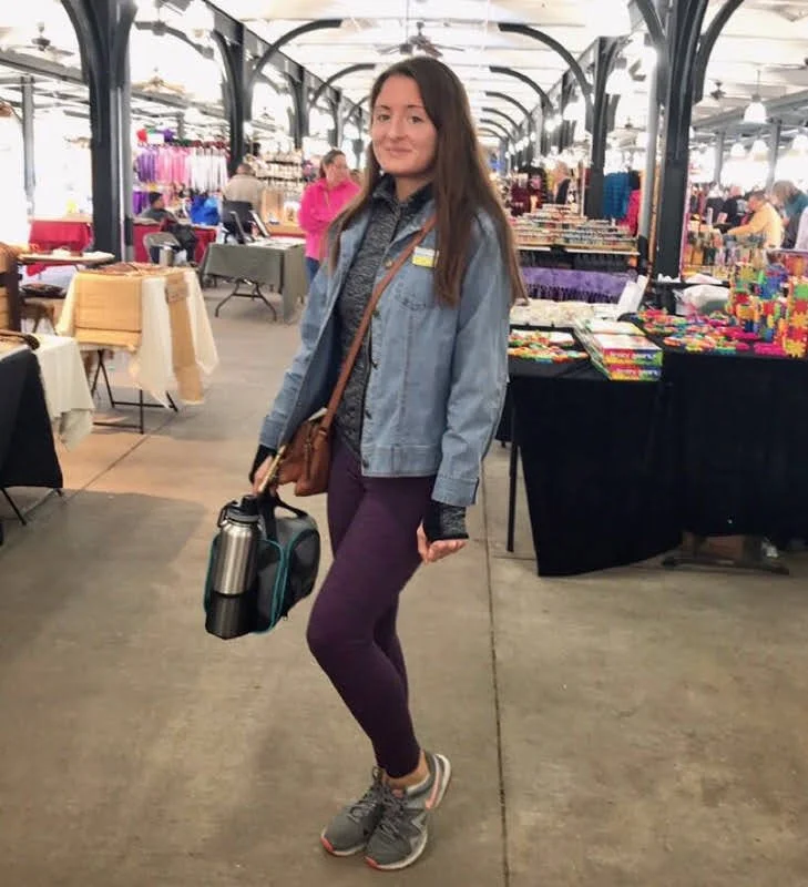 Jenny in denim jacket and leggings at an indoor market, holding thermos and lunch bag, surrounded by various stalls.