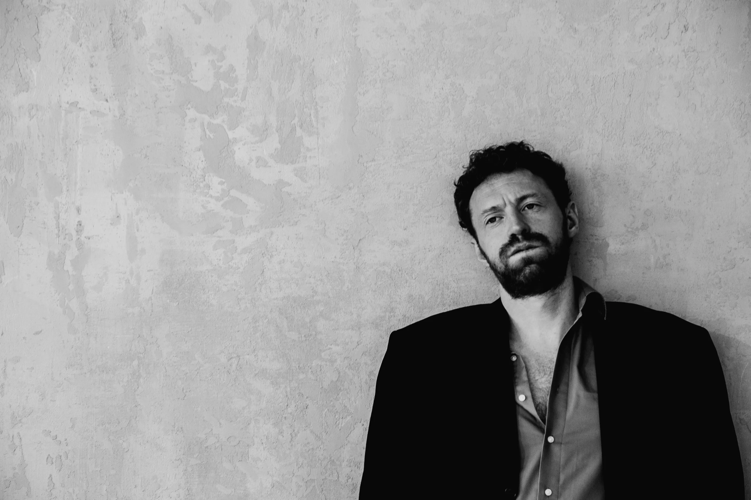 Black and white photo of a man with dark, curly hair and facial hair, wearing a dark blazer over an unbuttoned shirt, standing against a textured wall with a serious expression.