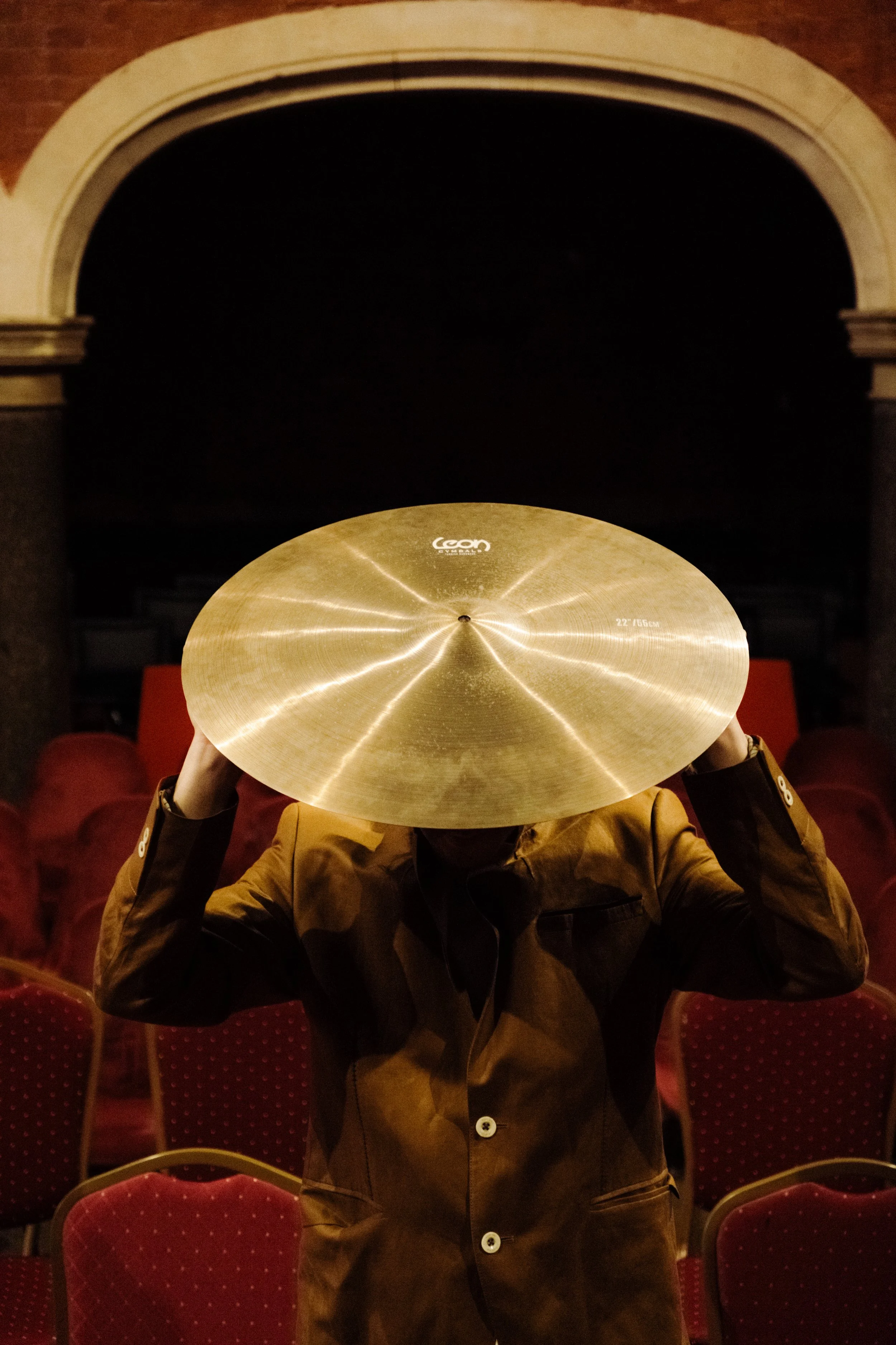 Person holding a shiny gold cymbal in front of their face in an auditorium with red chairs.