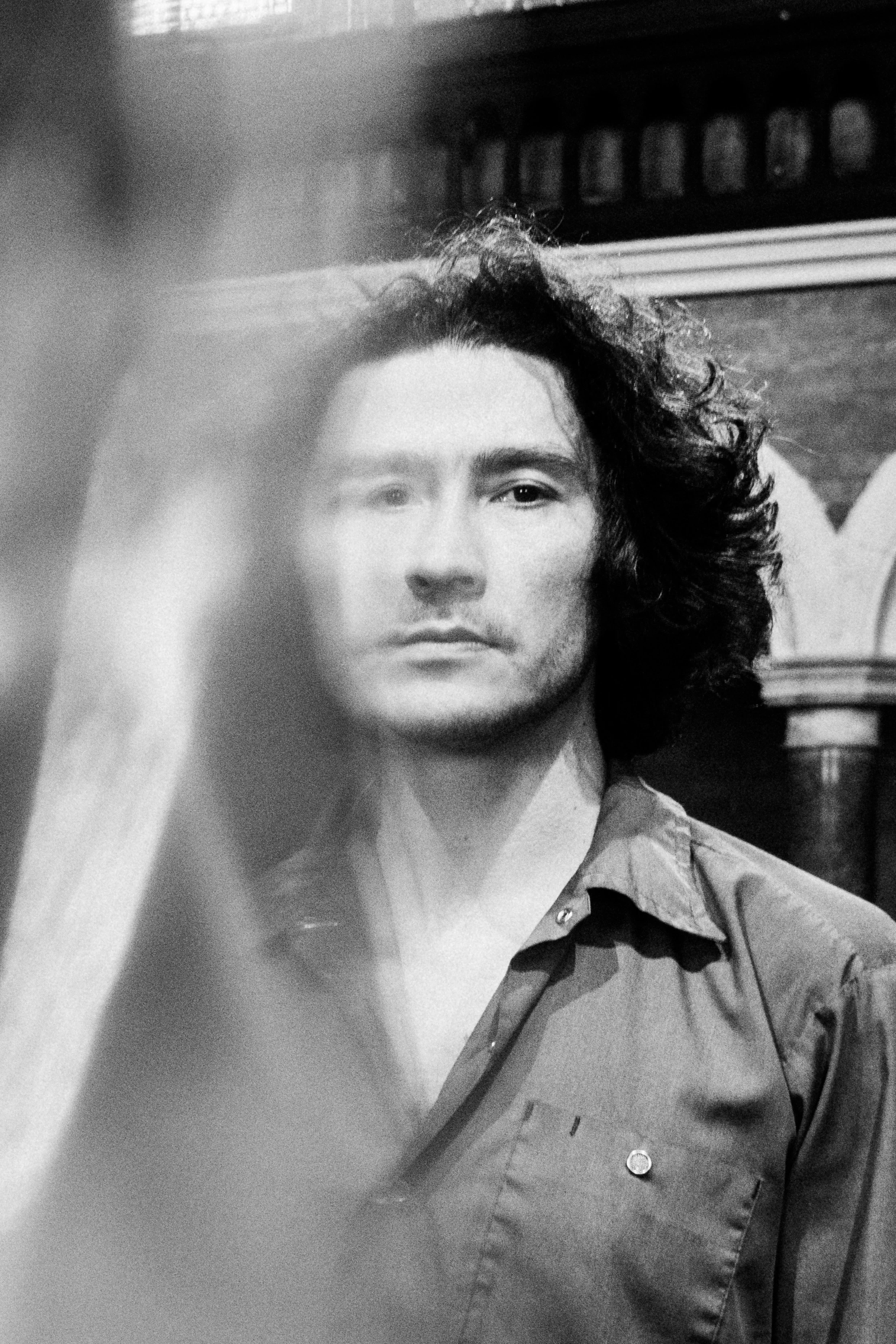 Black and white photo of a young man with long, curly hair, wearing a button-up shirt.