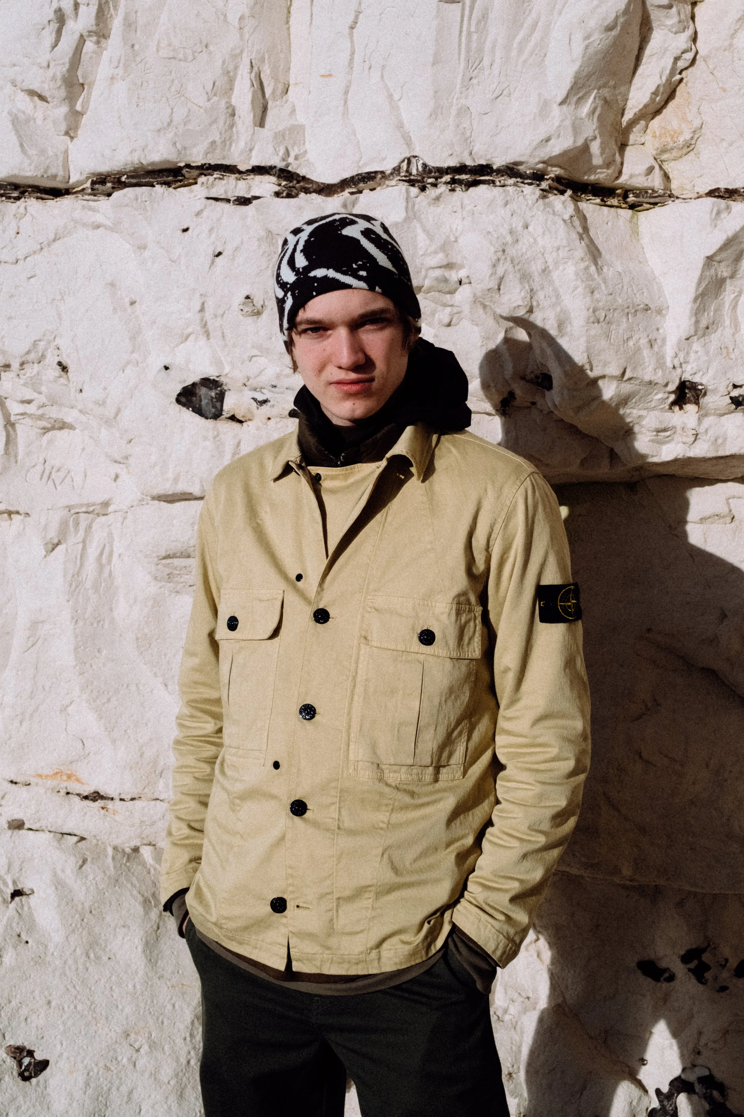 A young man wearing a beige jacket and a black and white patterned beanie standing in front of a white rocky wall.