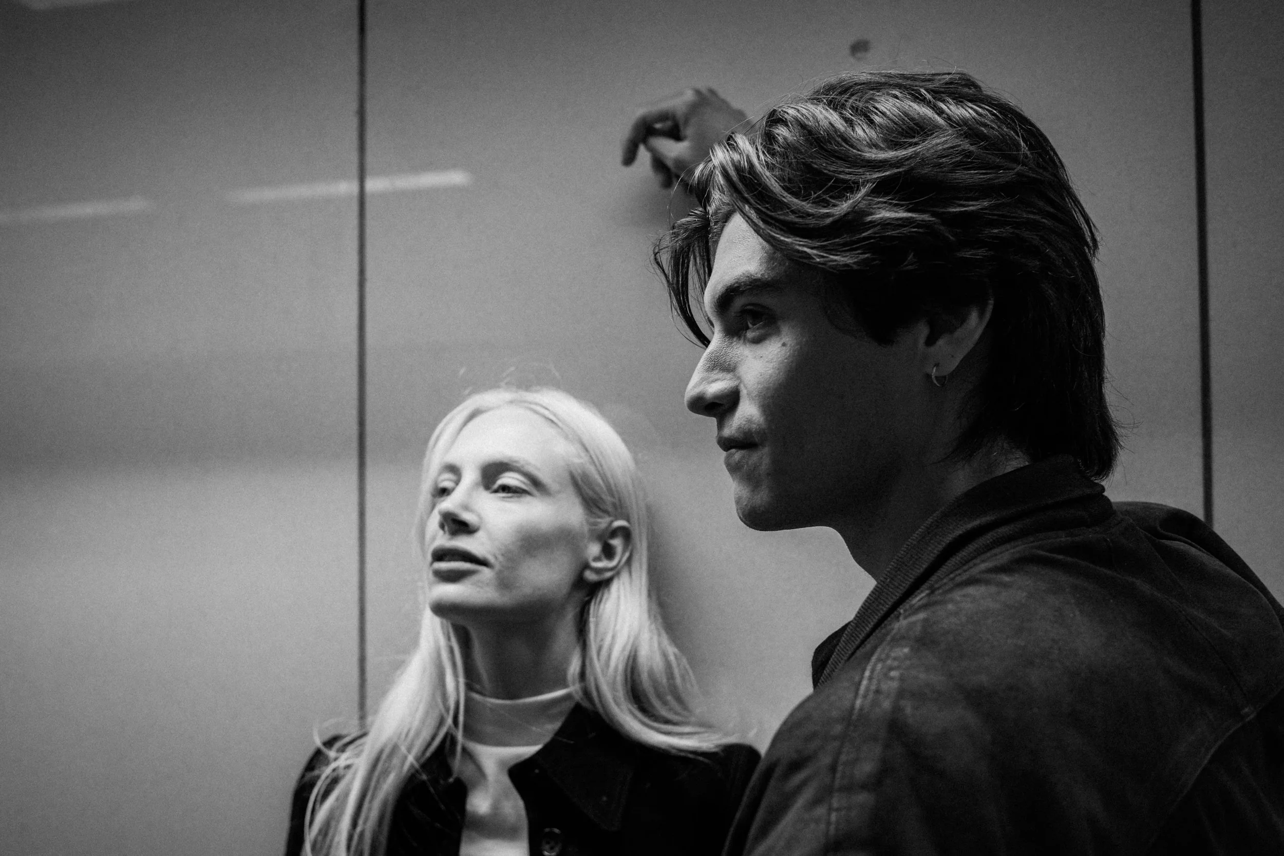 Black-and-white photo of a young woman with long blonde hair and a young man with dark, tousled hair, both standing in front of a mirror in a modern interior space. The woman appears to be looking off to the side with a slight smile, and the man is g