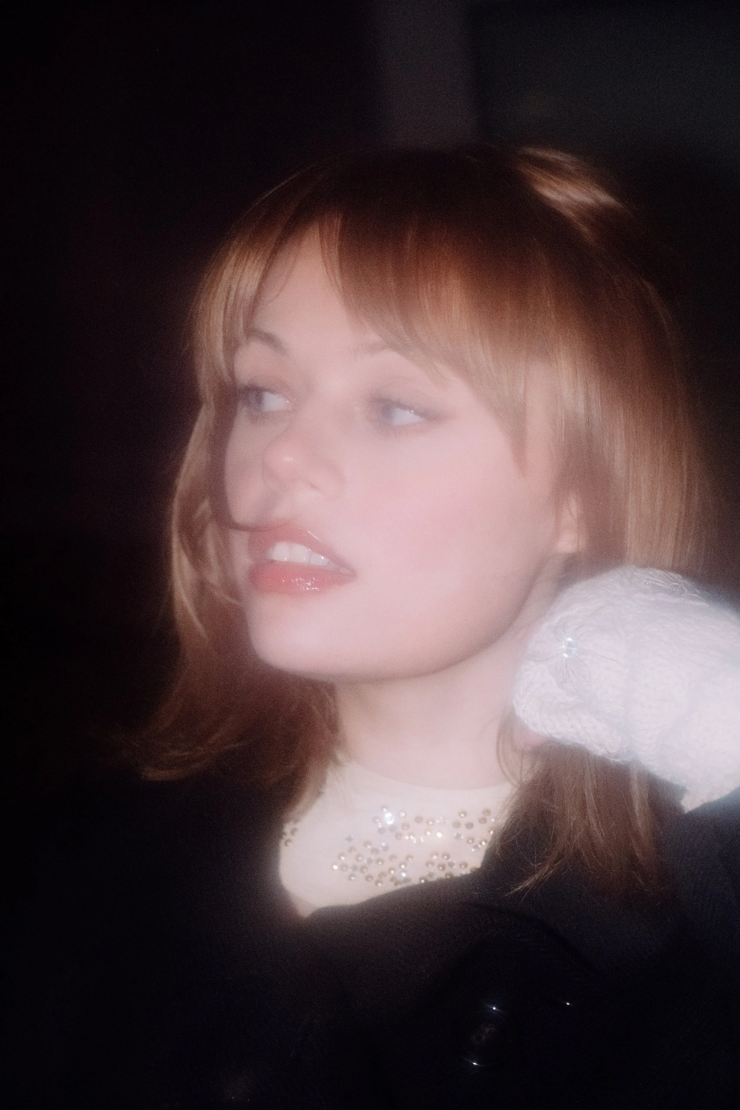 Close-up of a woman's face with light brown hair, smiling and looking to the side, wearing a white dress with embellishments.