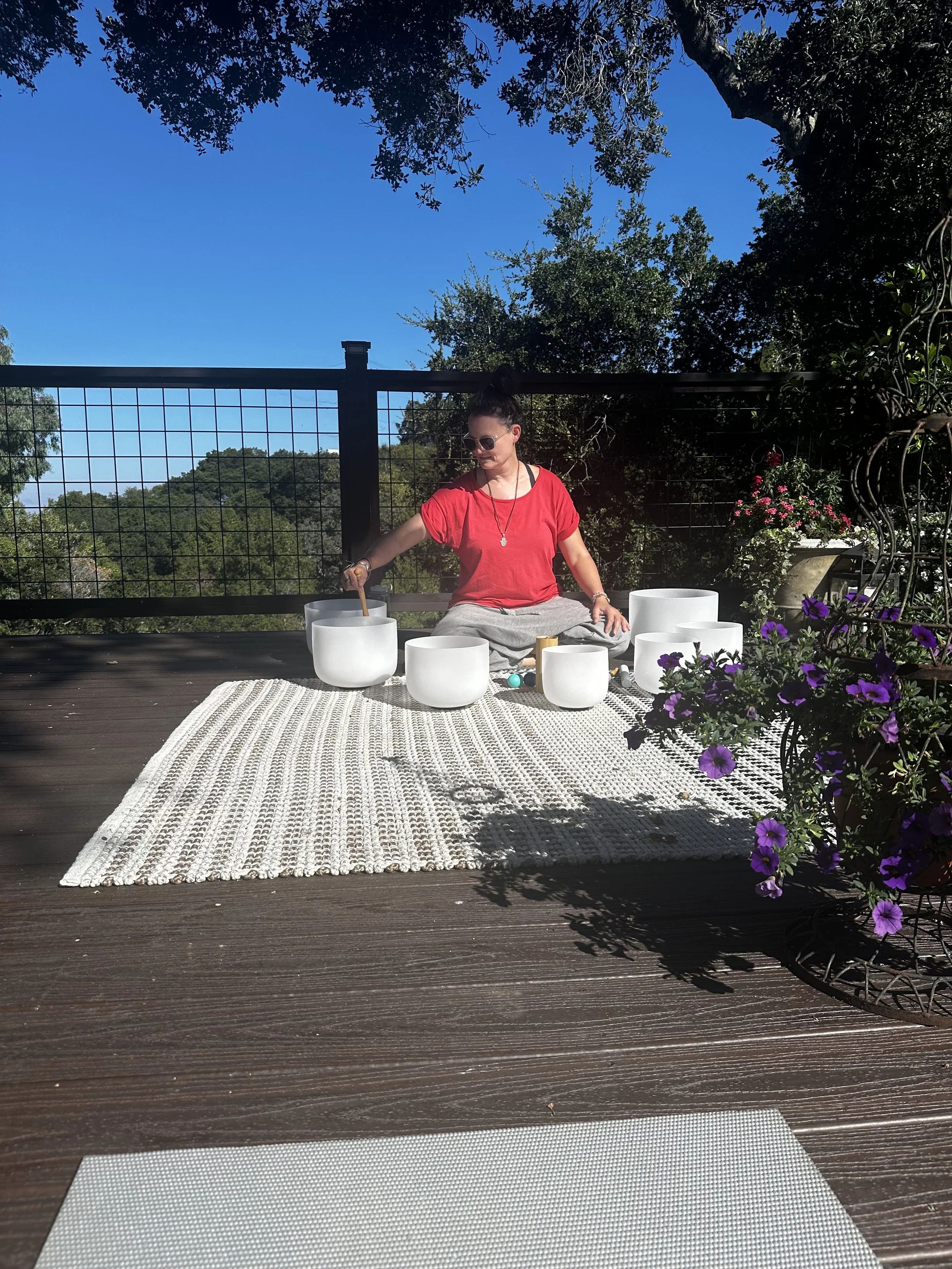 A lady playing crystal singing bowls outside on a deck with a blue sky and tree overhead.
