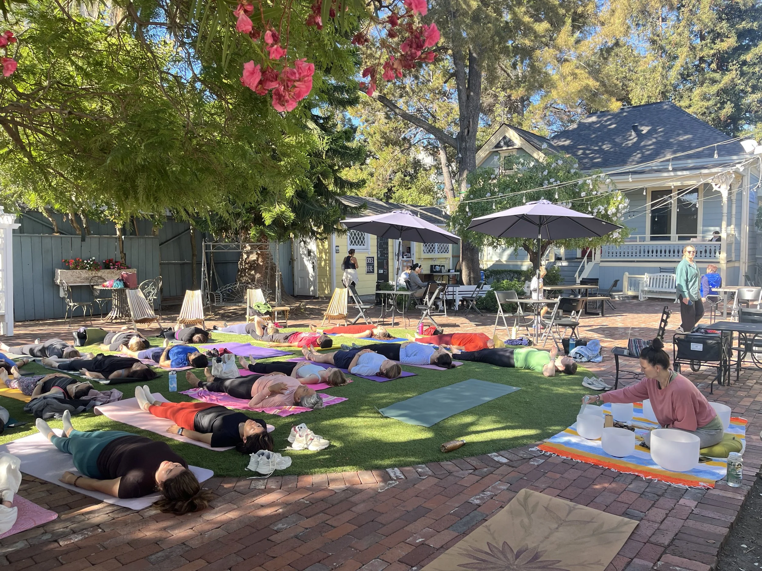 An outdoor setting at a coffee shop with a group of people lying on yoga mats under the trees while a woman plays crystal singing bowls.