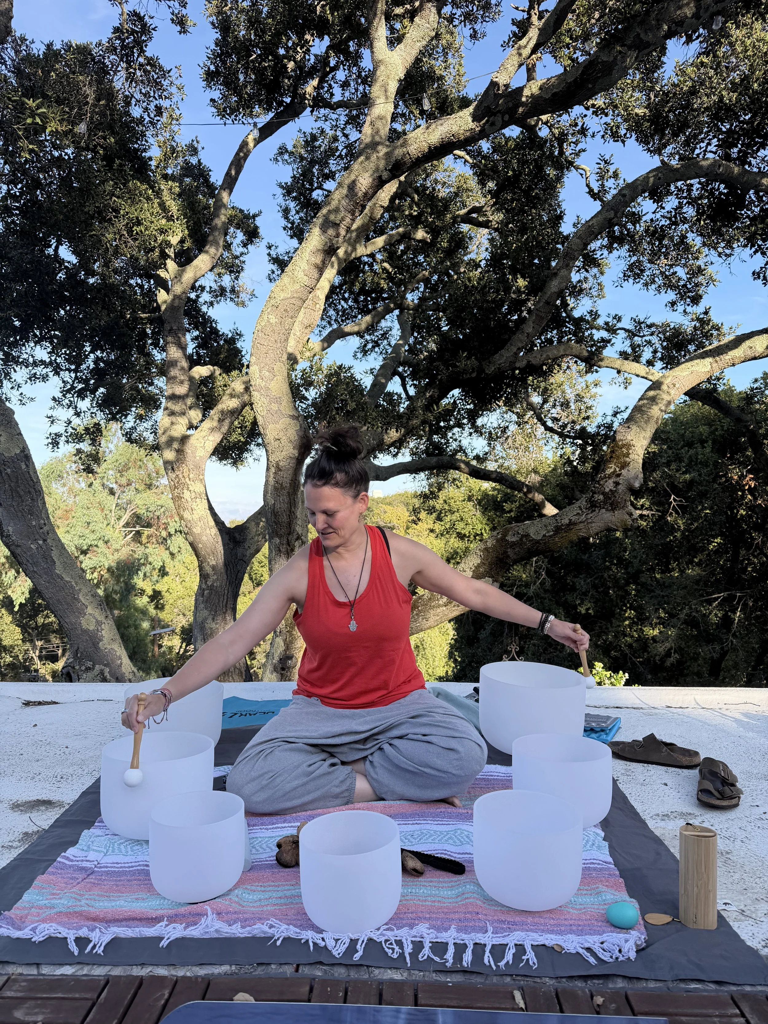 A woman playing crystal singing bowls wearing a red top and with a big tree and blue sky behind her.