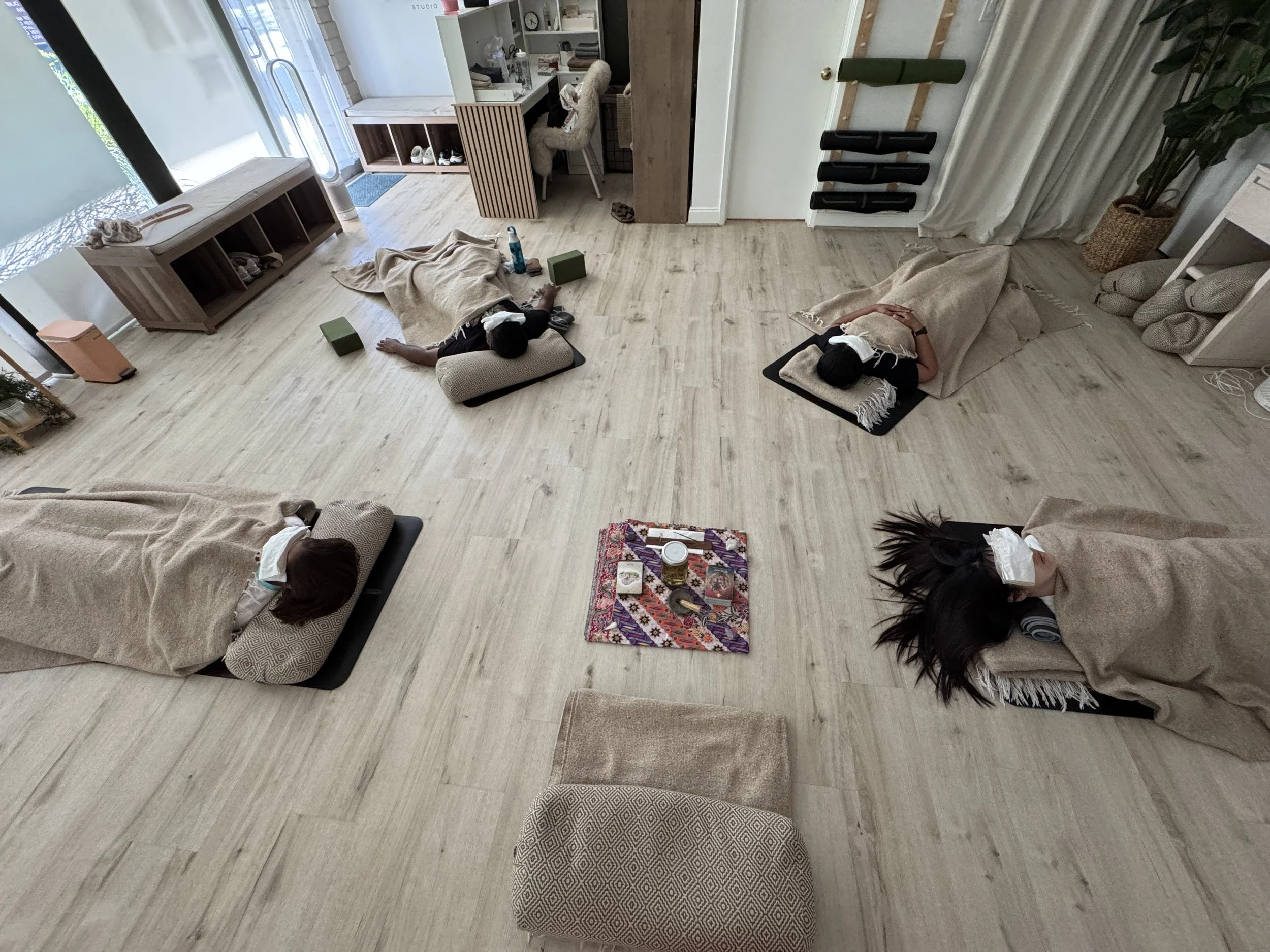 A group of people lying down in a circle on yoga mats, covered with blankets and with eye pillows while they meditate and receive reiki.