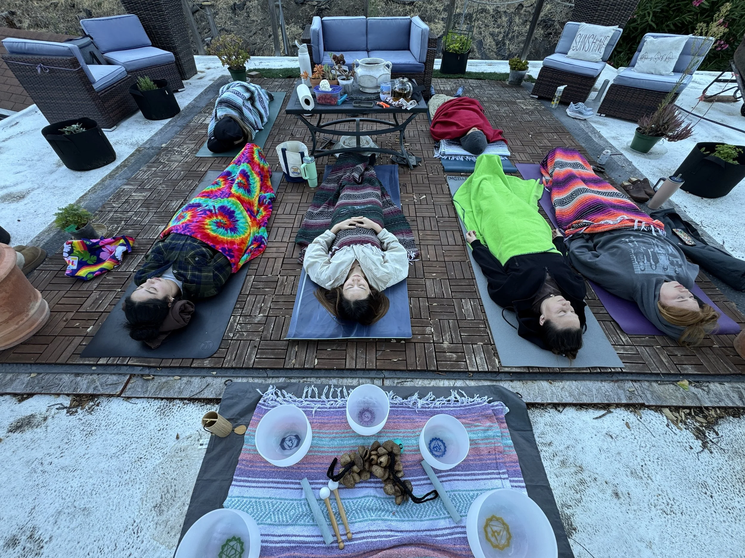 A group of people lying down with blankets on a wood platform with patio furniture in the background and crystal singing bowls in the forefront..