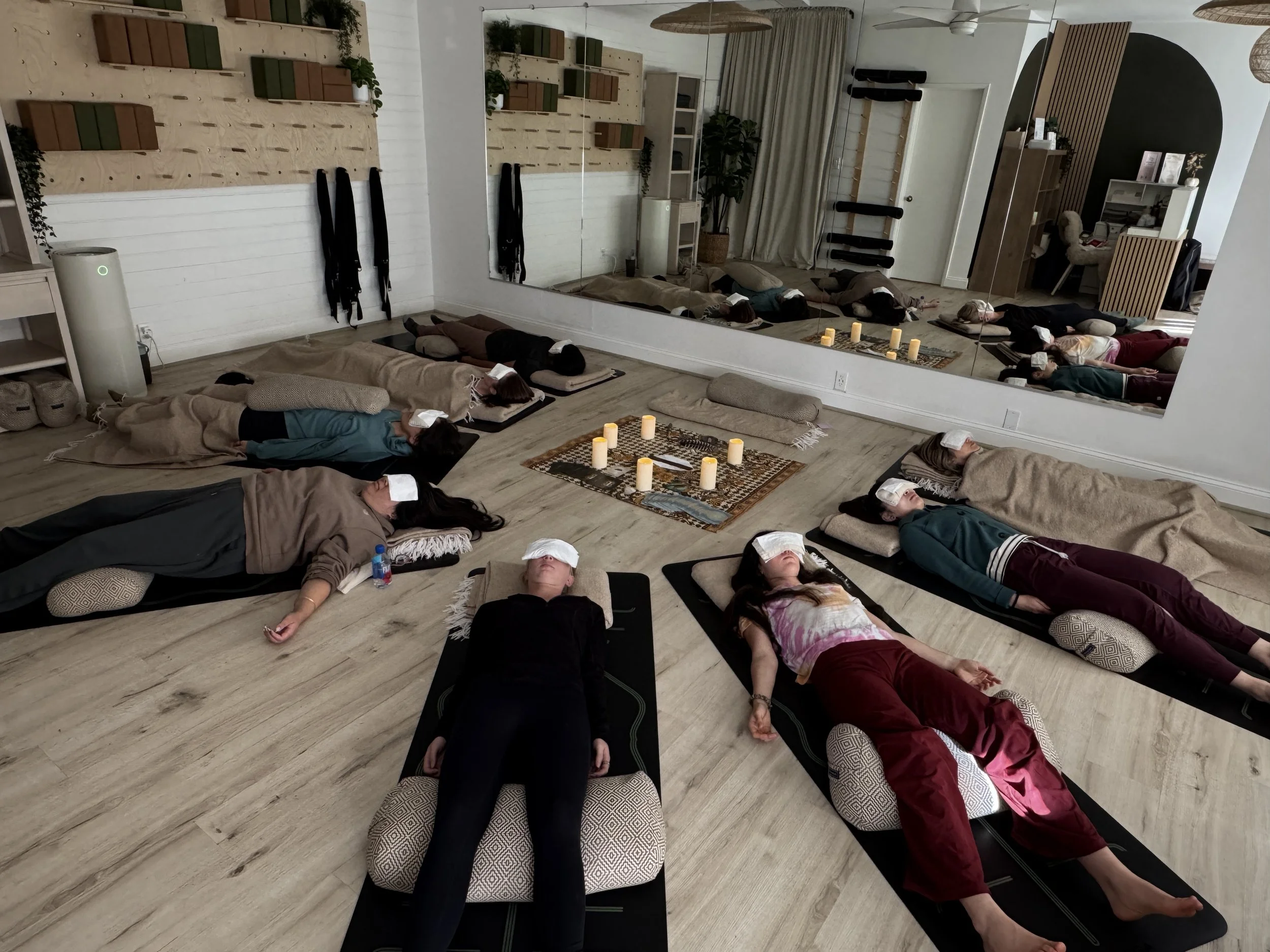 A group of people lying down in a circle on yoga mats, covered with blankets and with eye pillows while they meditate and receive reiki.