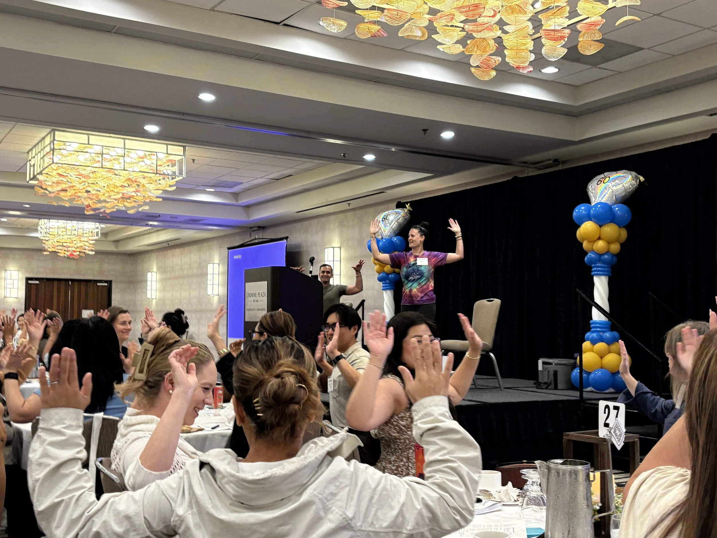 A woman standing on a stage in large room with a large group of people, leading them through some yoga poses.