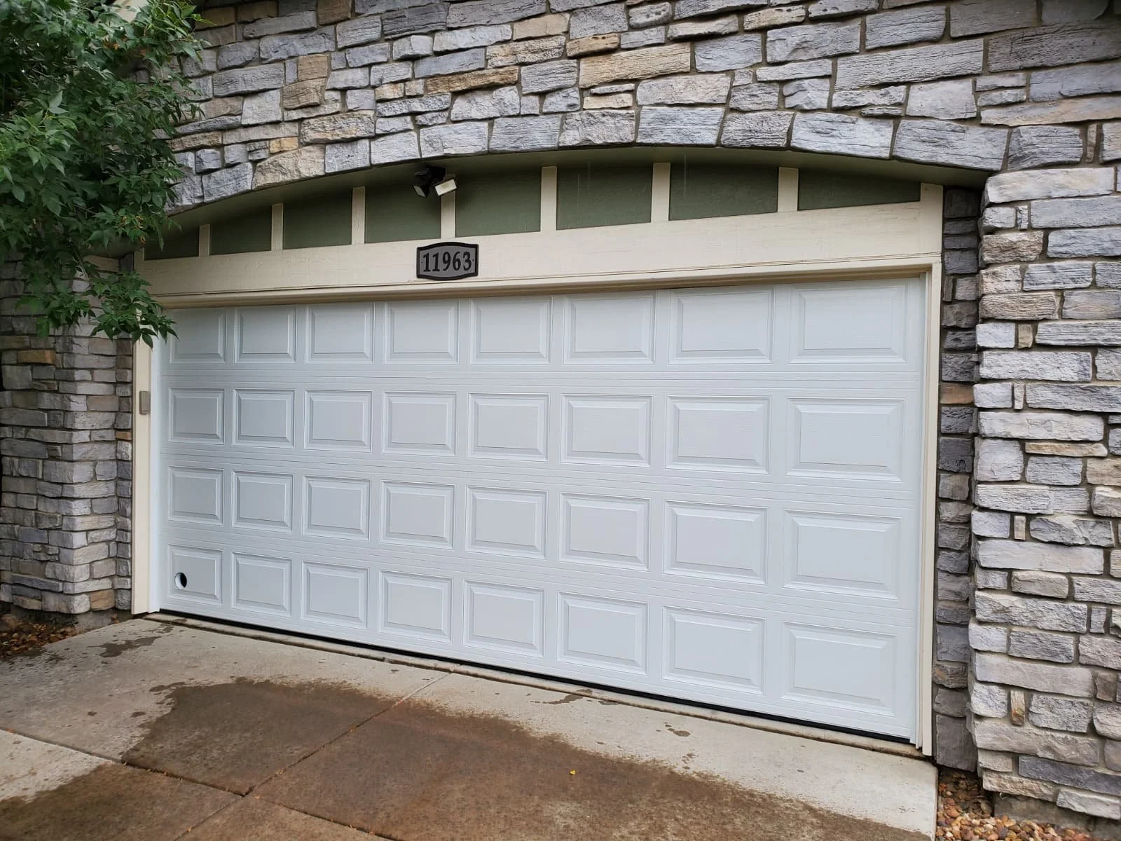 A white garage door with rectangular embossed panels, framed by stone wall siding, with a house number 11963 sign above, and a small black circular vent at the bottom left corner of the door.