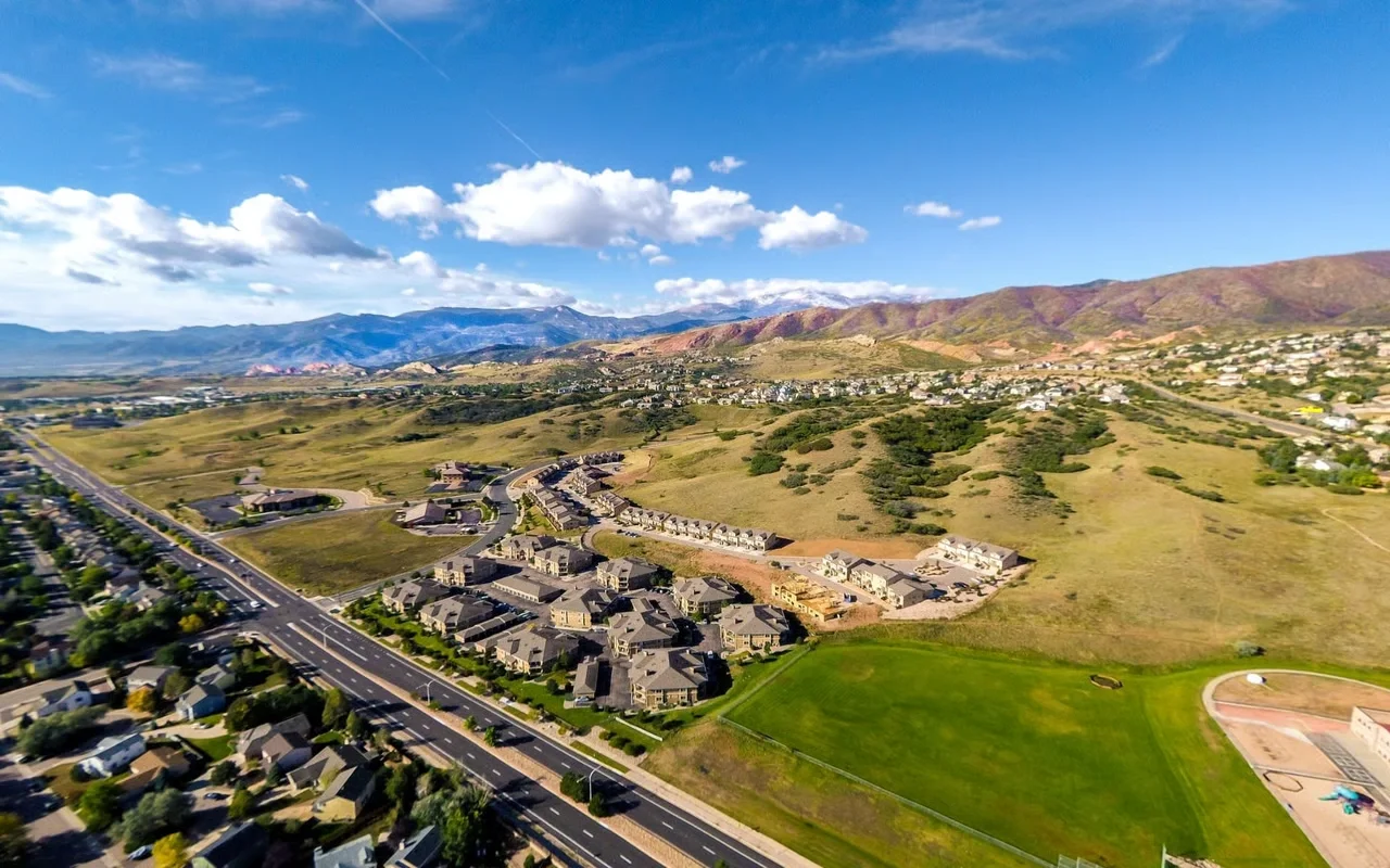 Aerial view of a suburban neighborhood with houses, a sports field, and surrounding mountains under a partly cloudy sky.