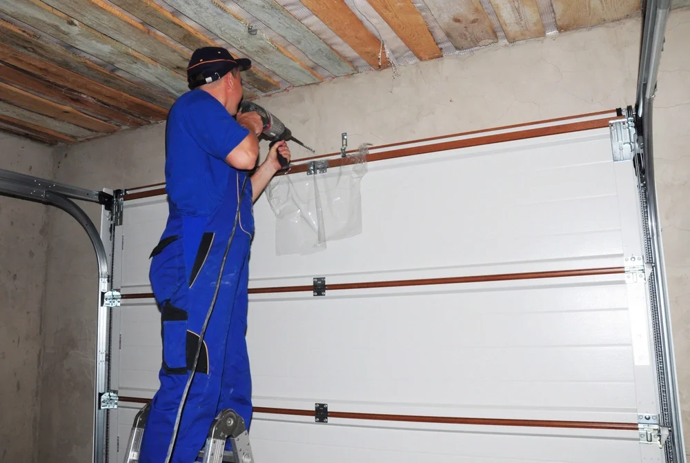 A person in blue coveralls and a black cap is installing or repairing a garage door with a power drill in a garage with a concrete wall and wooden ceiling.