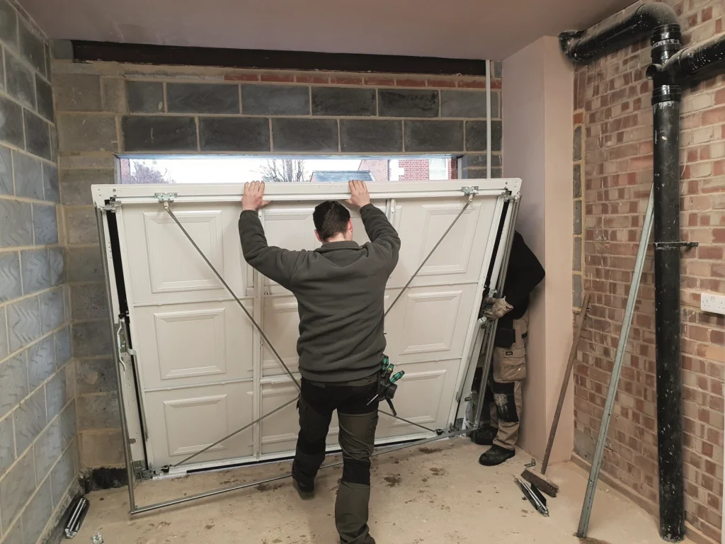 Two workers are installing or repairing a white garage door inside a room with exposed brick and concrete block walls.