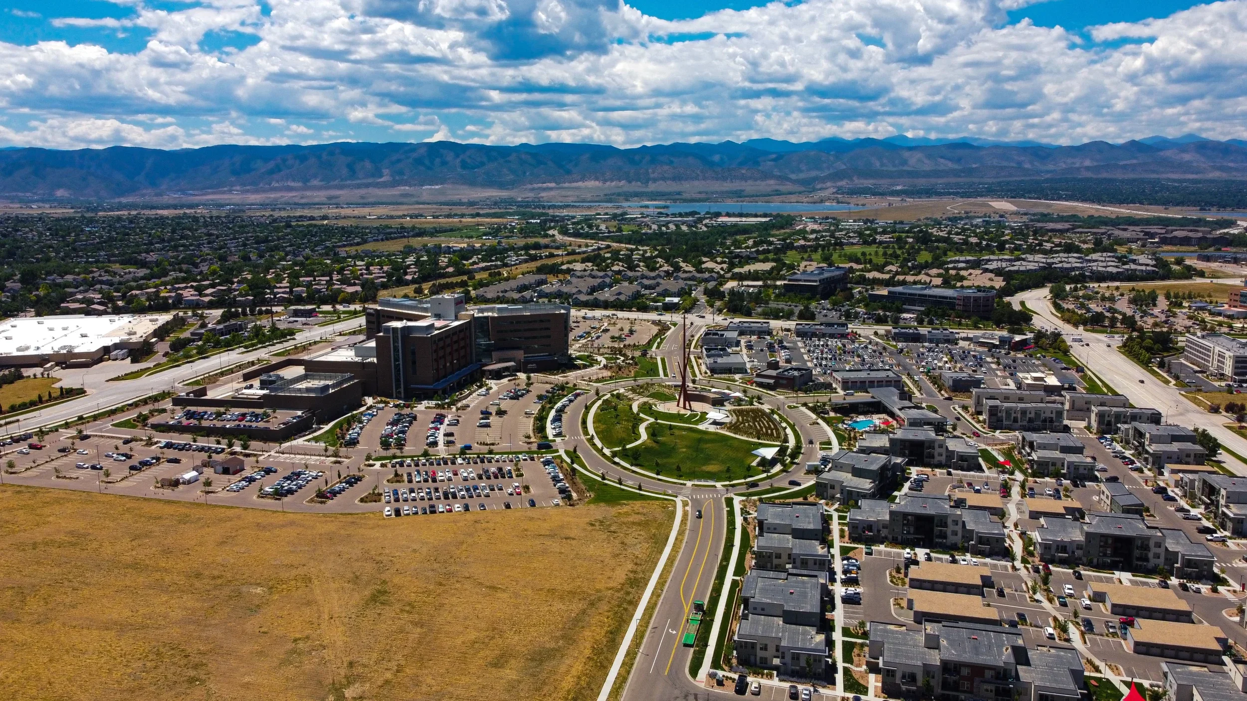 Aerial view of a developed urban area with a circular park at the center, surrounded by parking lots, roads, and various buildings, with mountains and a river in the background under a partly cloudy sky.
