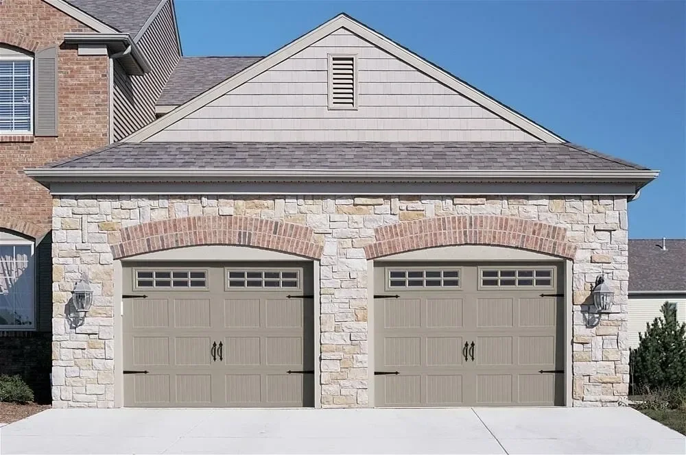 Front view of a two-car garage with stone and siding exterior, two tan garage doors with windows at the top, and outdoor lighting on each side. The house features a peaked roof with shingles and a clear blue sky.