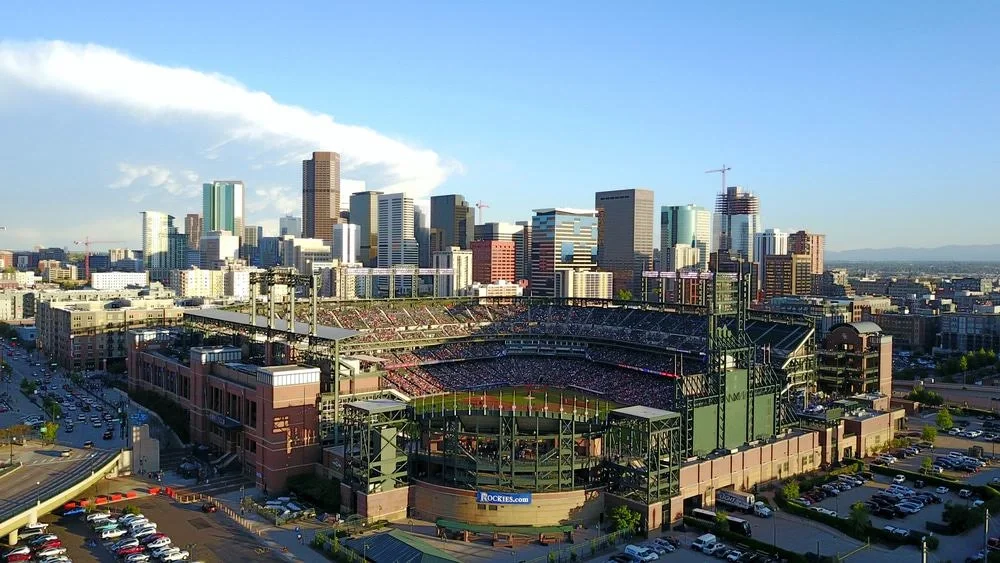 Aerial view of a baseball stadium in a city with tall skyscrapers in the background, filled with spectators during game day.