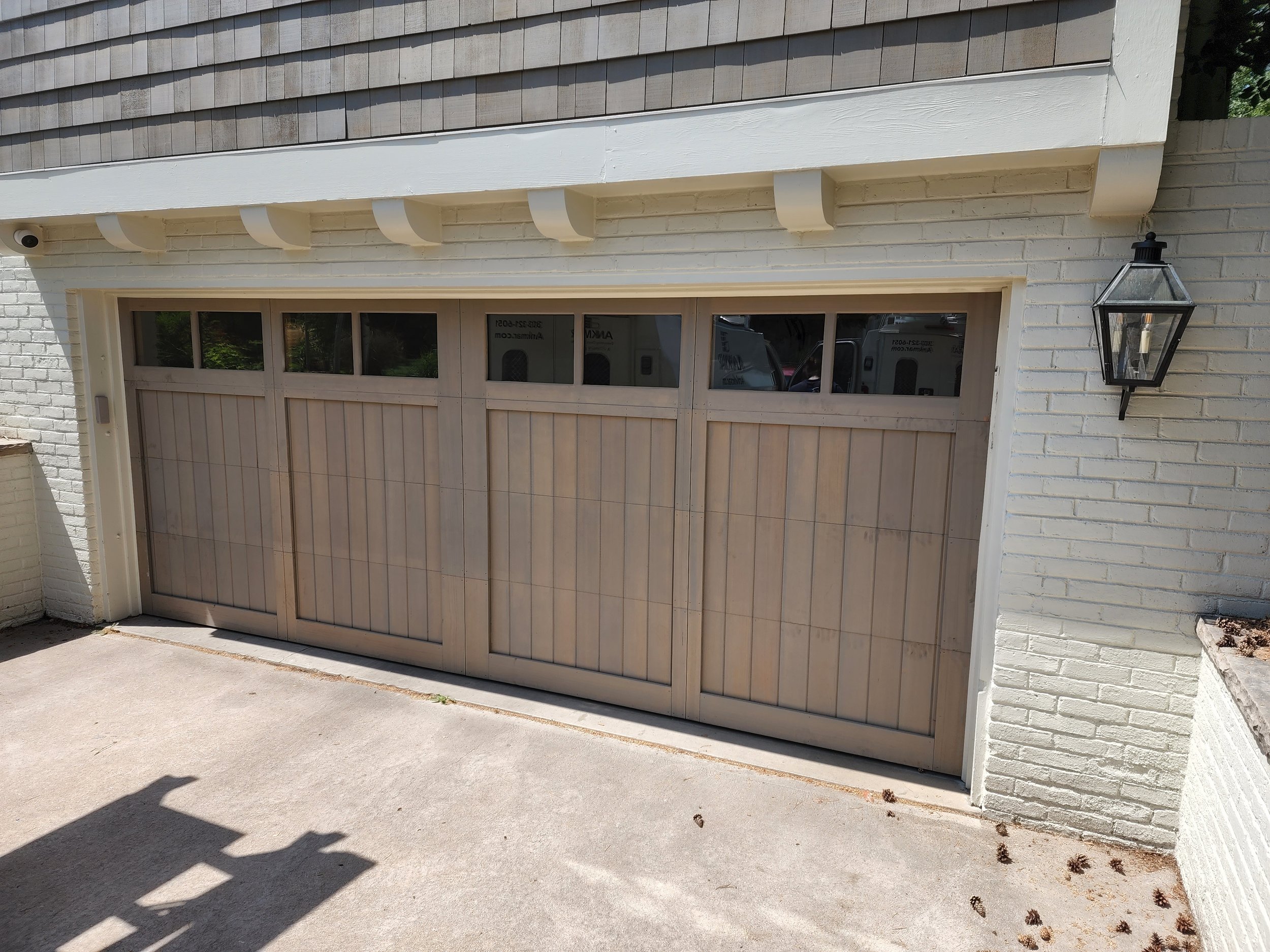 A beige garage door with small windows at the top, set in a white brick house with a black outdoor lantern on the right side.