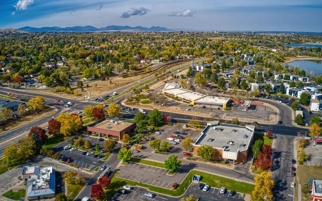 Aerial view of a suburban area showing roads, residential neighborhoods, commercial buildings, parking lots, and natural scenery including trees and lakes, during daytime with partly cloudy sky.