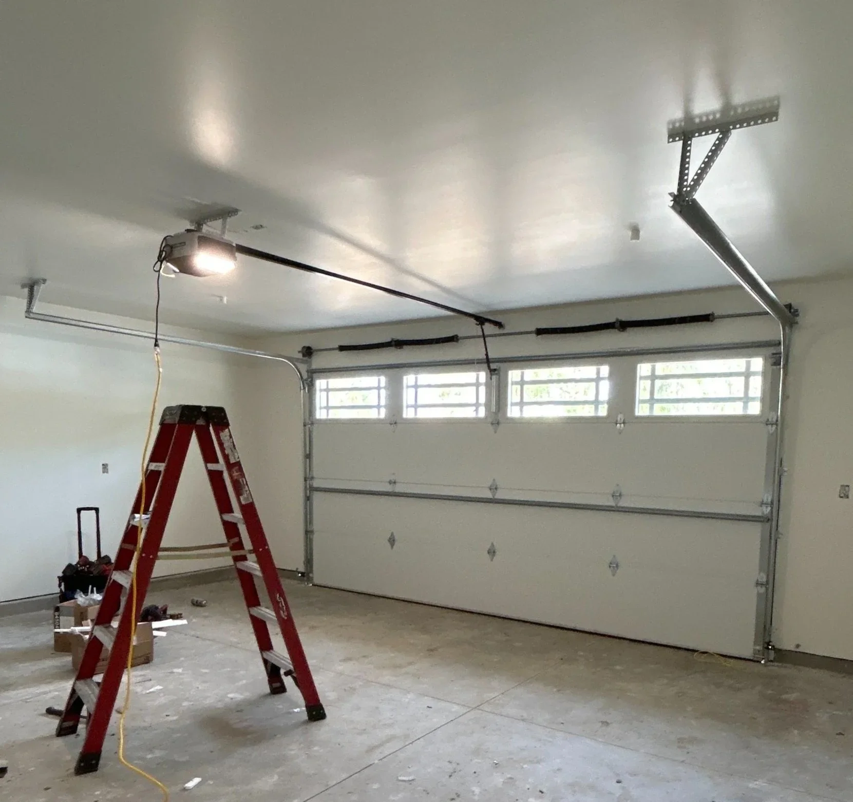 A garage with an automatic garage door, a red step ladder, and construction tools. The ceiling has an electric garage door opener and tracks. The garage floor is unfinished concrete.