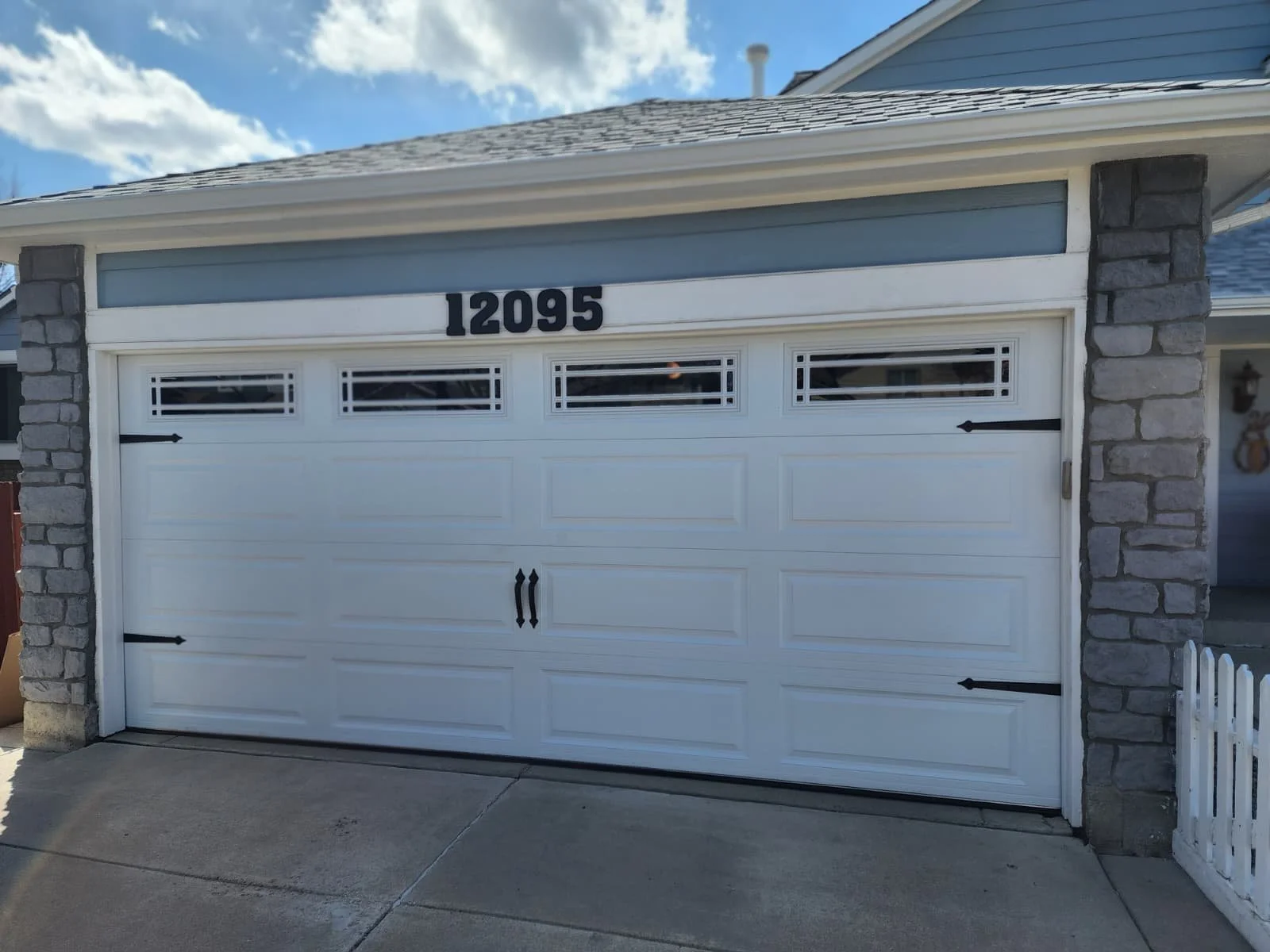 White garage door with no windows, black house number 12095 above, side stone columns, small white picket fence on right, concrete driveway in front, partly cloudy sky.