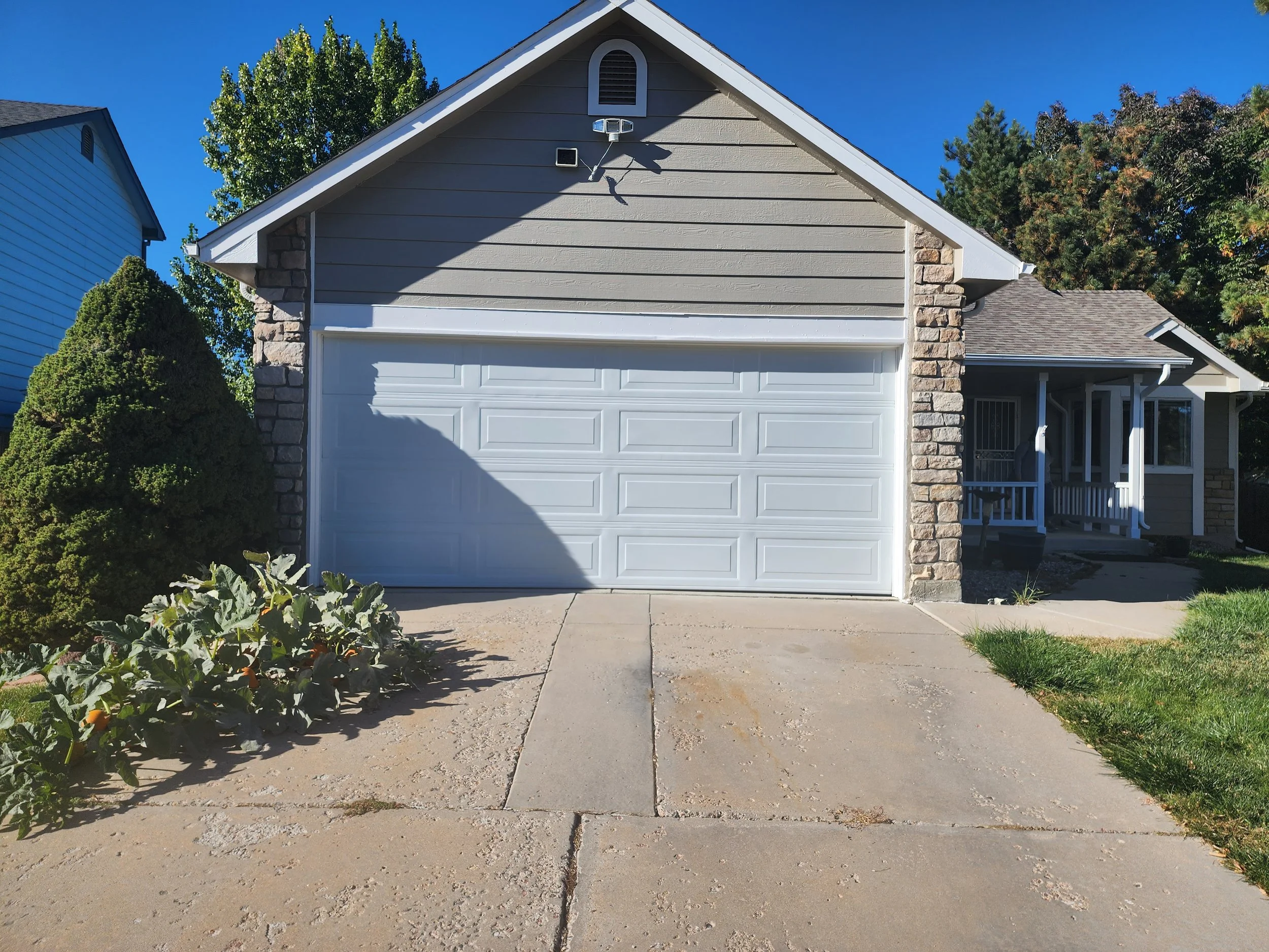 Front view of a suburban house with a white garage door, beige siding, rock accents, and a front porch with white railings. There is a driveway, some green grass, and plants on the left side. The sky is clear and blue.