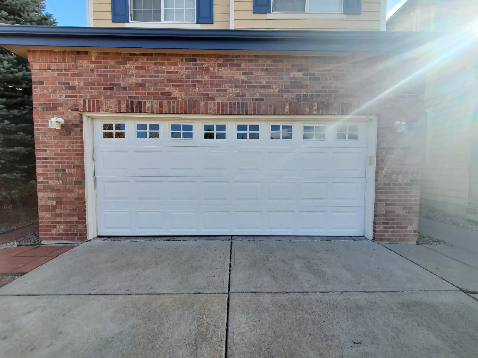 A white garage door with small windows at the top, set in a brick house with a concrete driveway in front, and two external wall-mounted lights on either side of the garage.