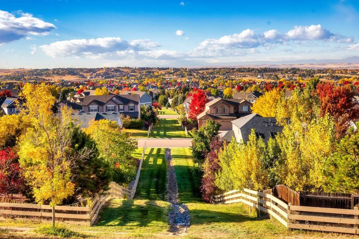 A residential neighborhood during fall with trees displaying yellow, orange, and red leaves. The houses are visible in the background under a blue sky with scattered clouds, and there is a green grassy area with a narrow drainage ditch running through it.